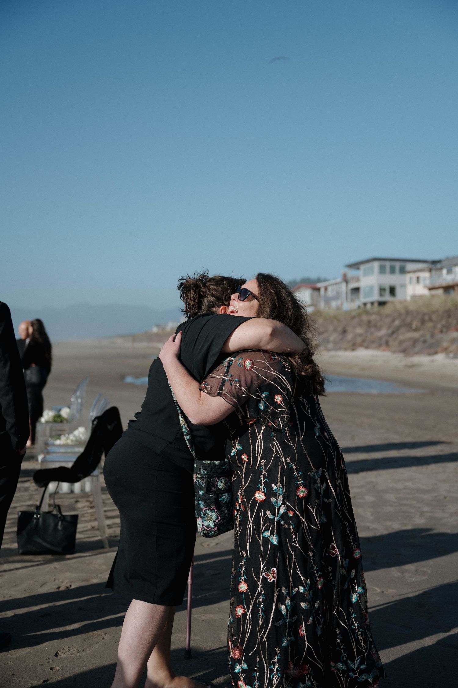 Two women share a warm embrace at an outdoor beach wedding celebration under a clear blue sky.
