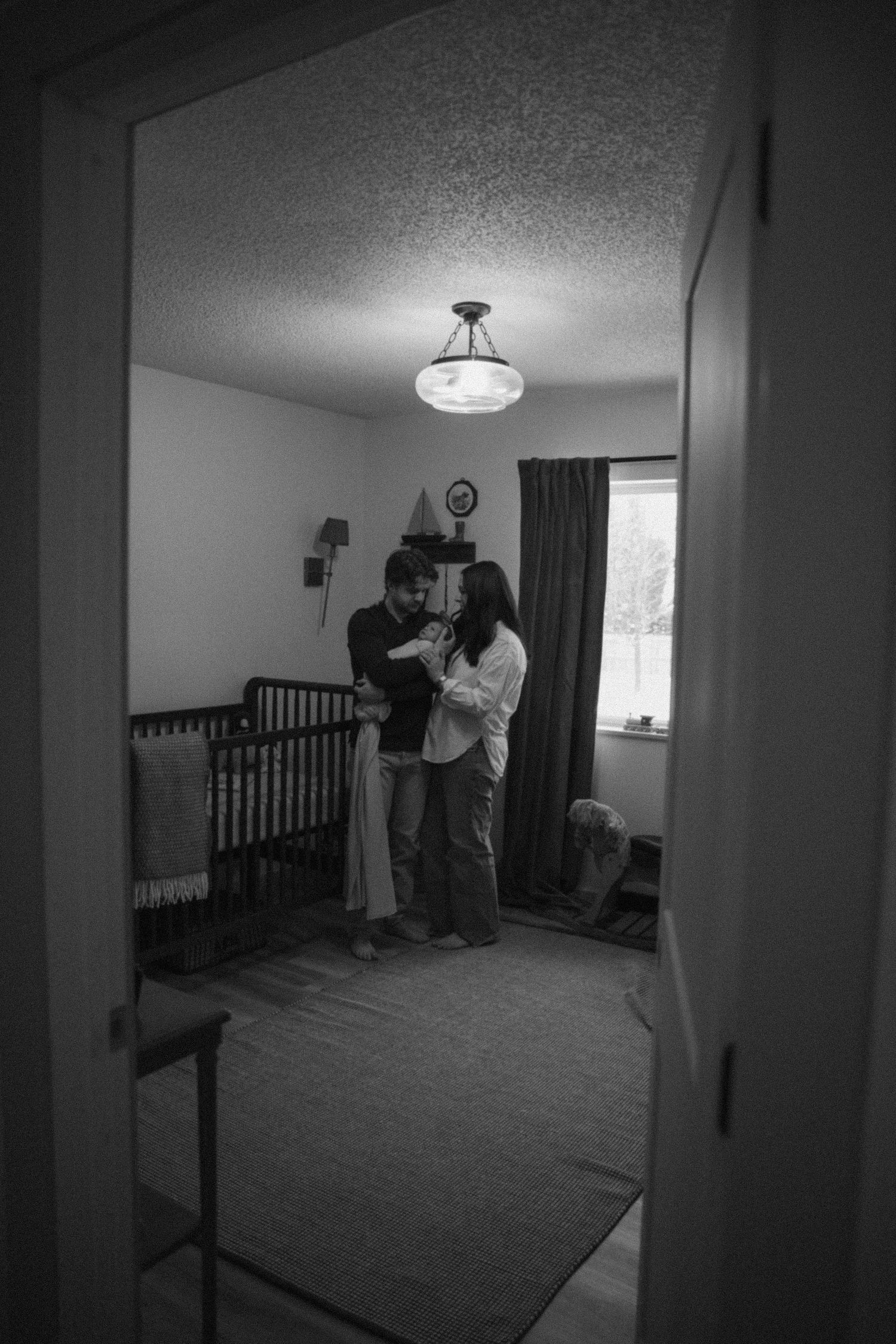 Black and white photo of new parents with newborn in nursery with wooden crib.