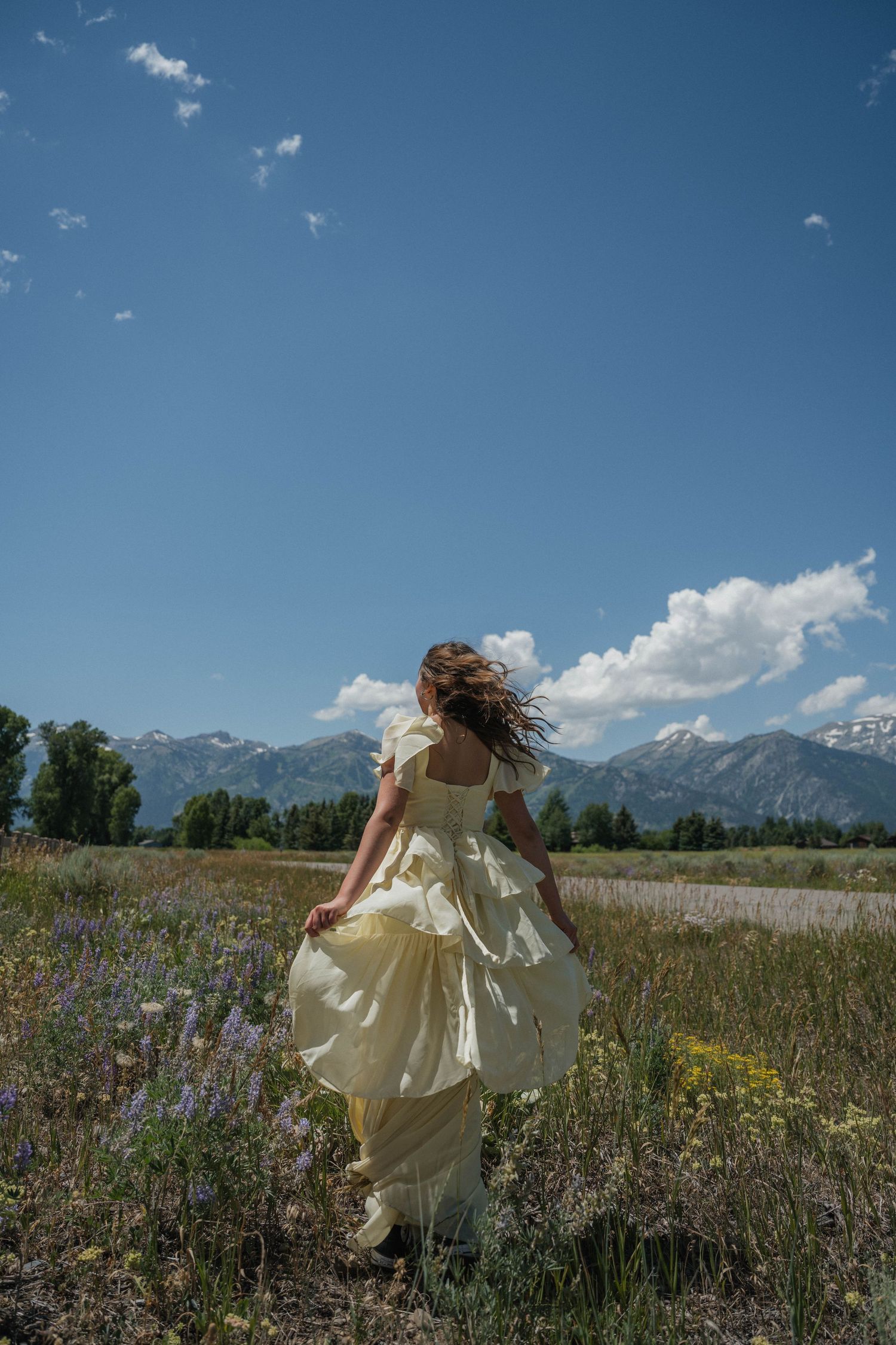 Woman in billowing yellow dress walking through wildflower field with wind-swept hair and Teton mountains.
