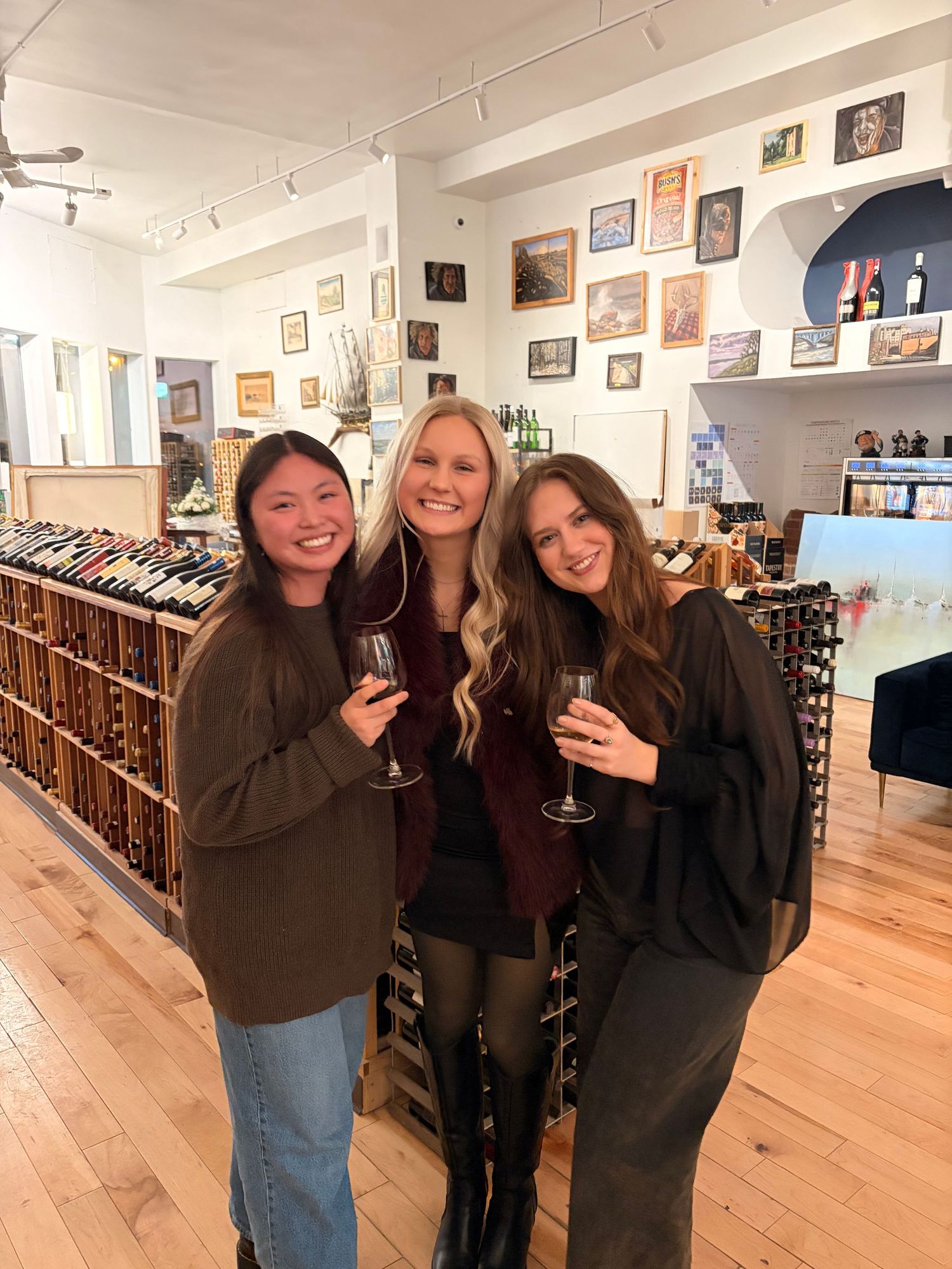 Three women smiling and holding wine glasses in a boutique wine shop with wood floors.