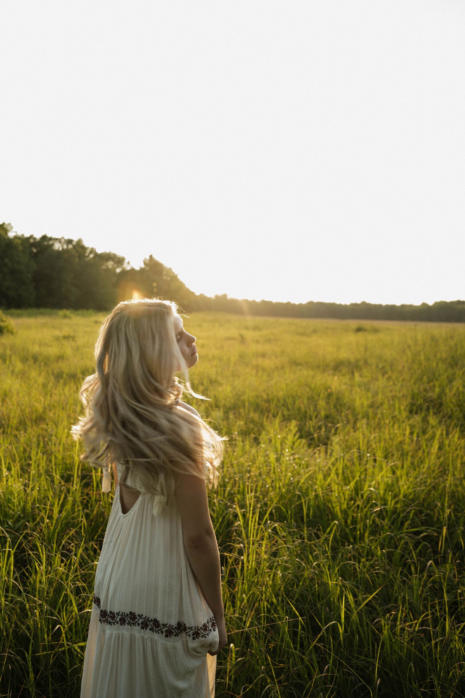 A blonde woman in a white dress stands in a sunlit green field with golden backlight at sunset.