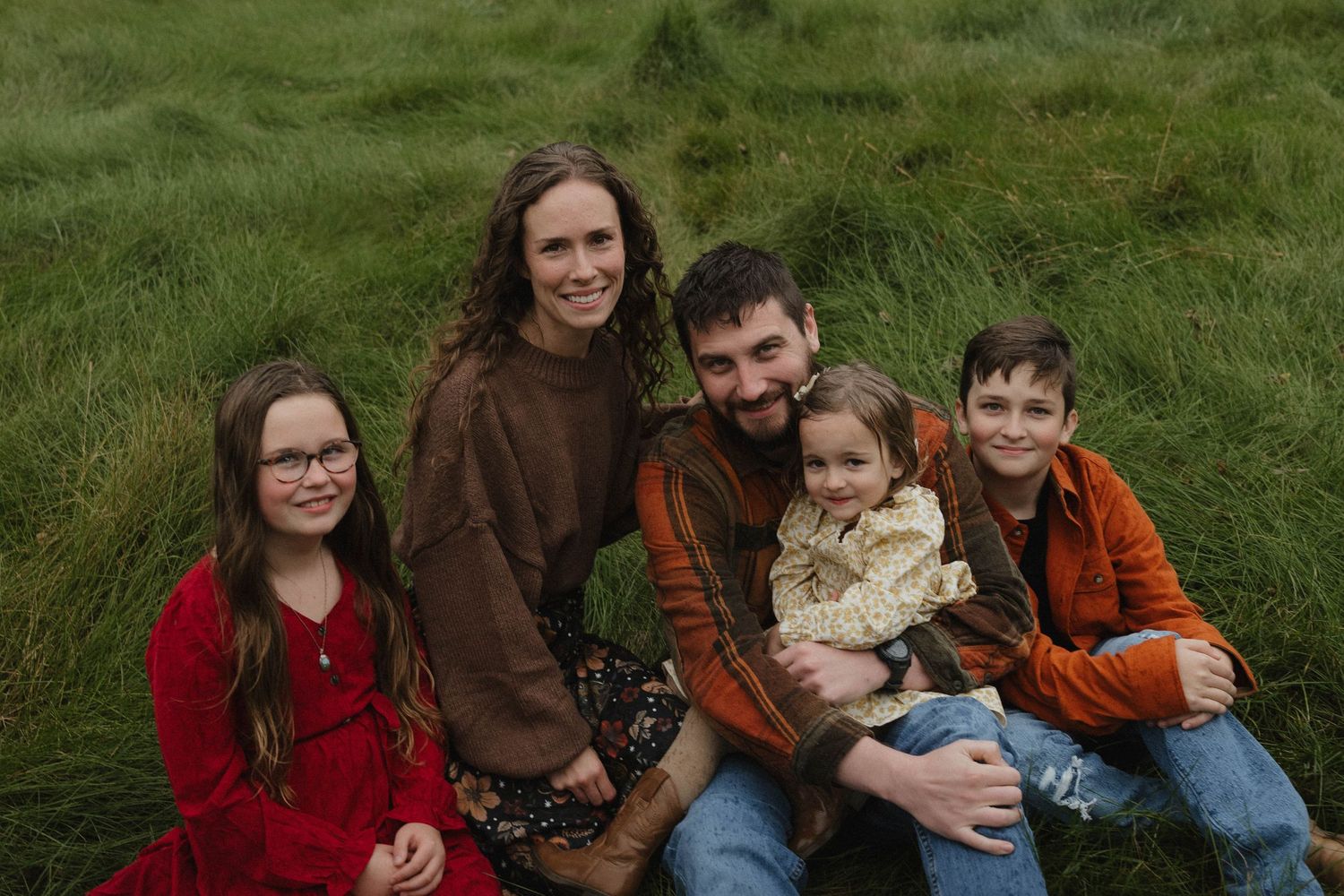 A happy family of five sitting together in a green grassy field during a fall photo session.
