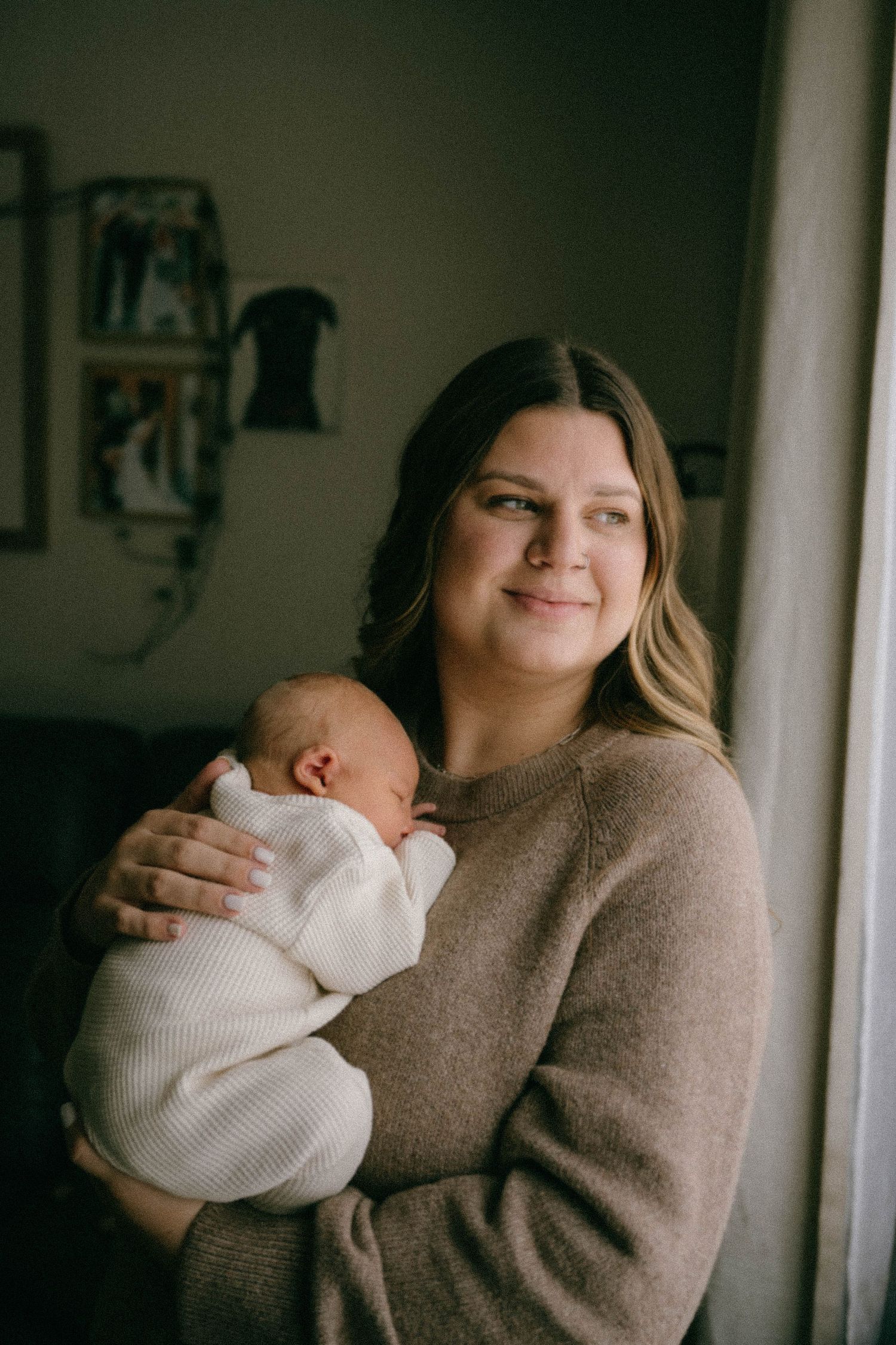 Smiling brunette mother in tan sweater holding sleeping newborn baby near window.