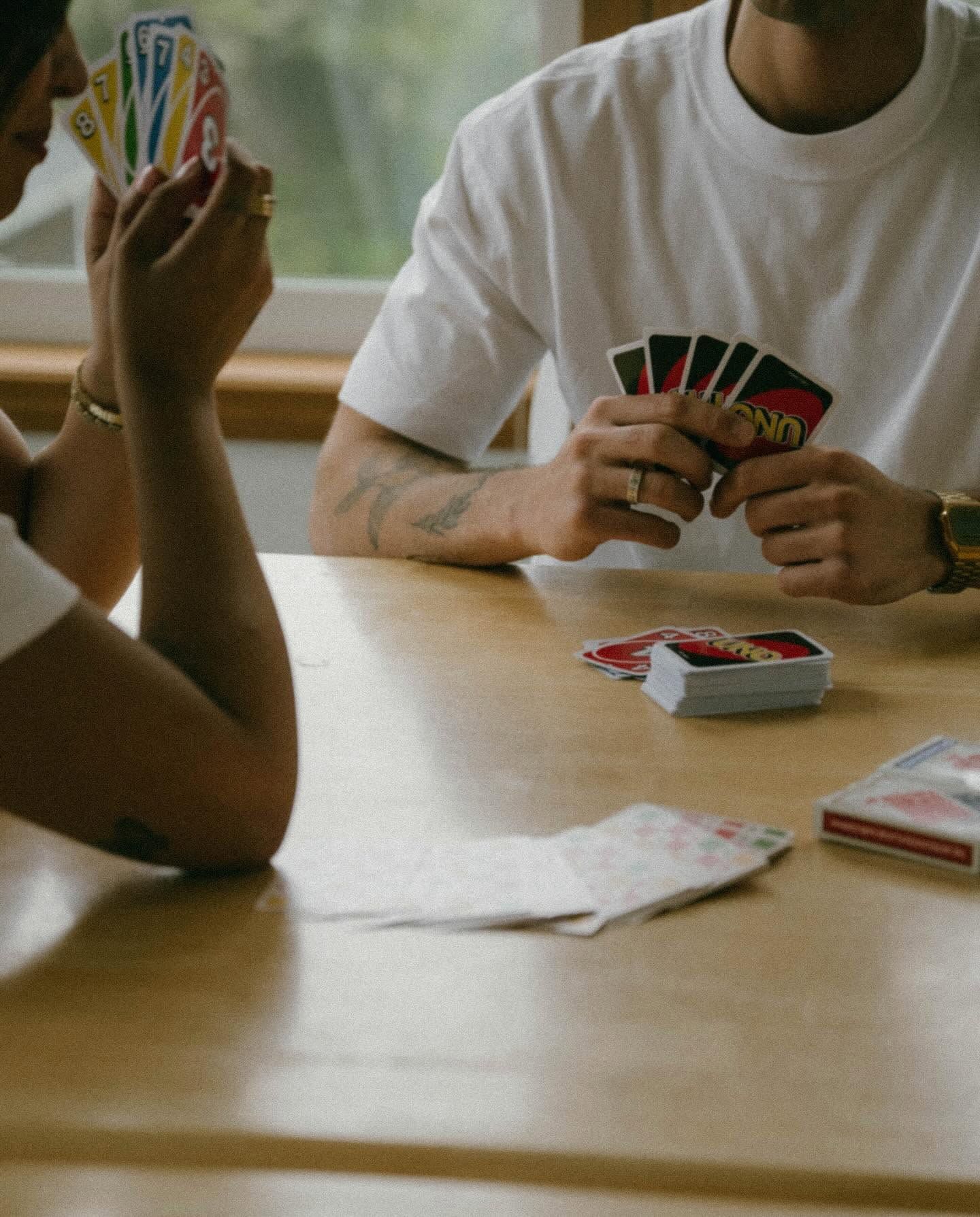Two people in white t-shirts playing Uno card game at a wooden table, holding colorful cards.