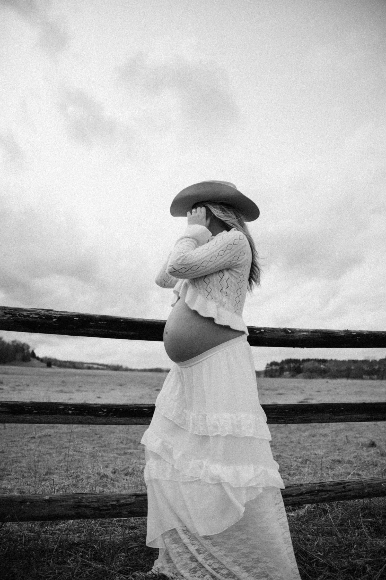 Black and white photo of pregnant woman in cowboy hat and white dress embracing her belly by a fence.