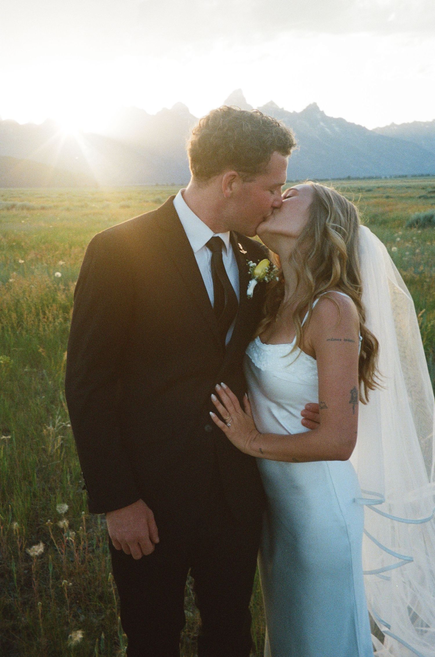 Bride and groom share romantic sunset kiss in meadow with Grand Teton mountains glowing behind them.