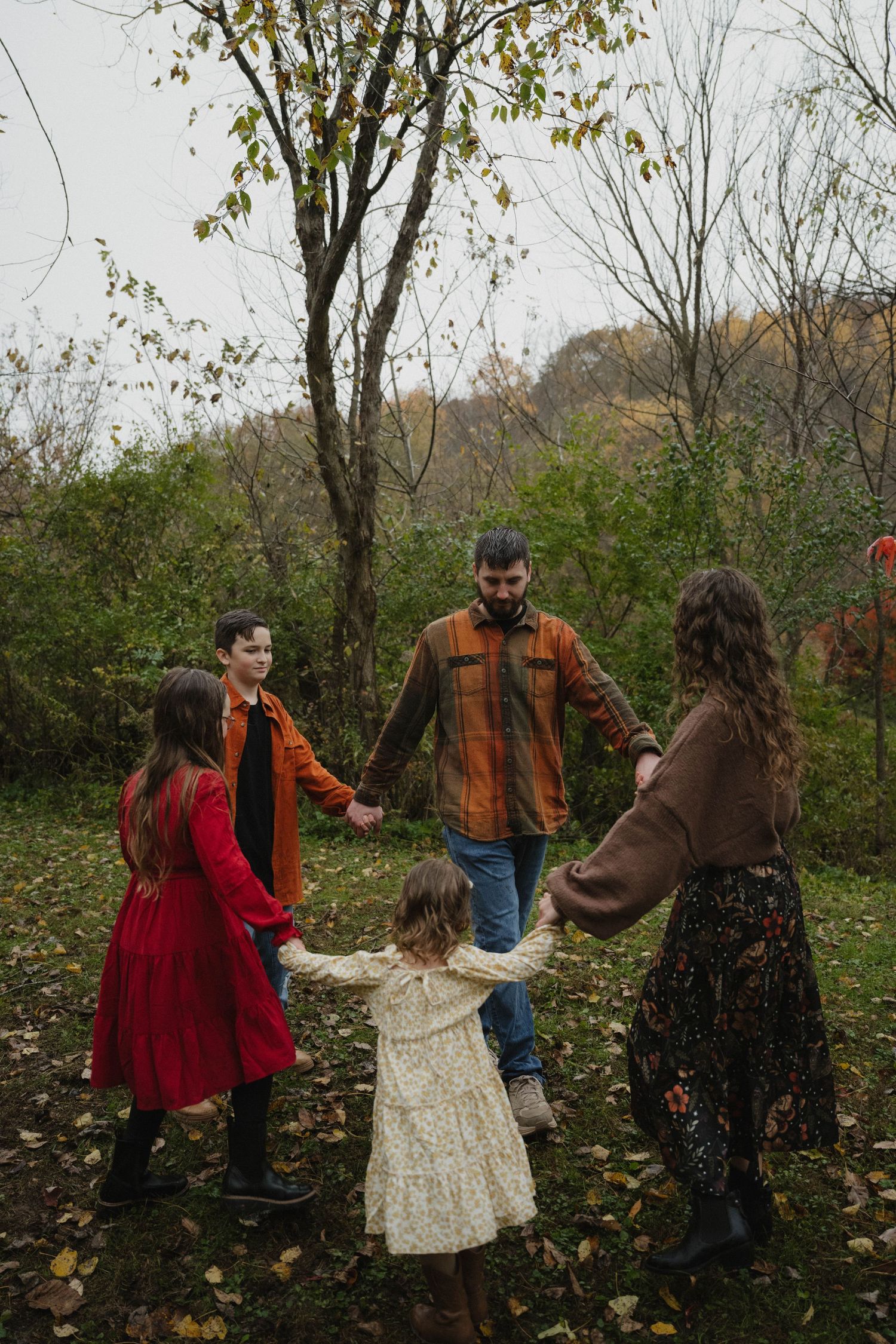 A family of five holding hands in a circle among colorful autumn trees and fallen leaves.