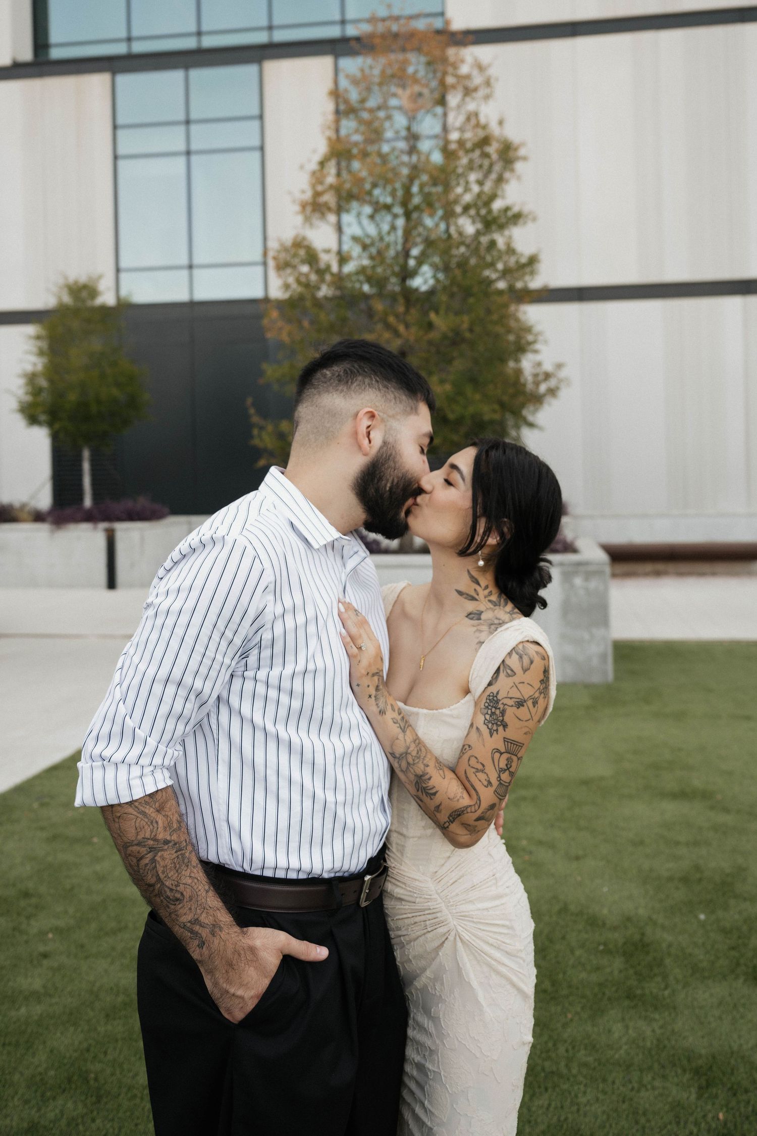 Tattooed couple sharing a kiss outdoors in wedding attire in front of a modern building.