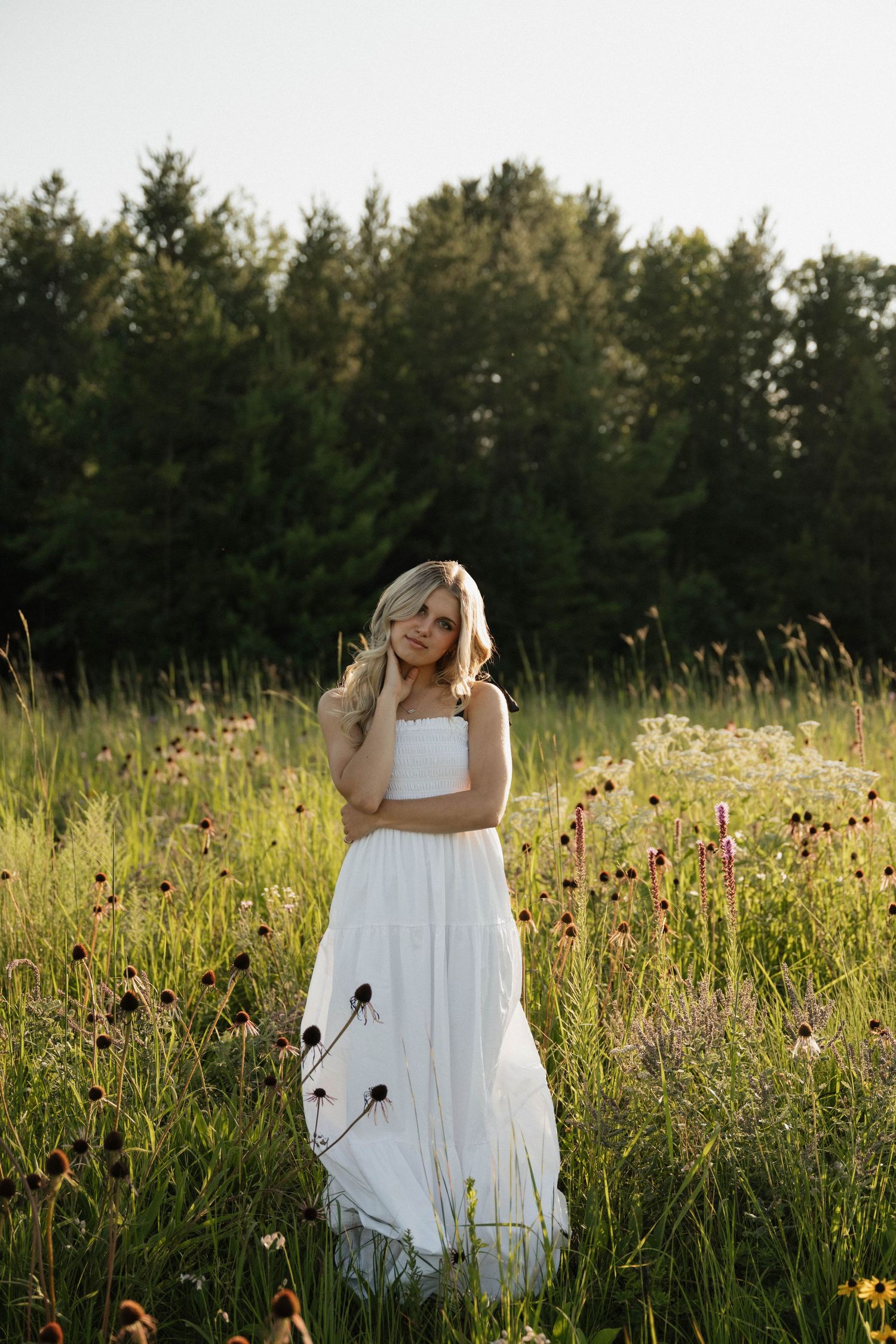 A blonde woman in a white dress stands in a sunlit wildflower meadow with trees behind.