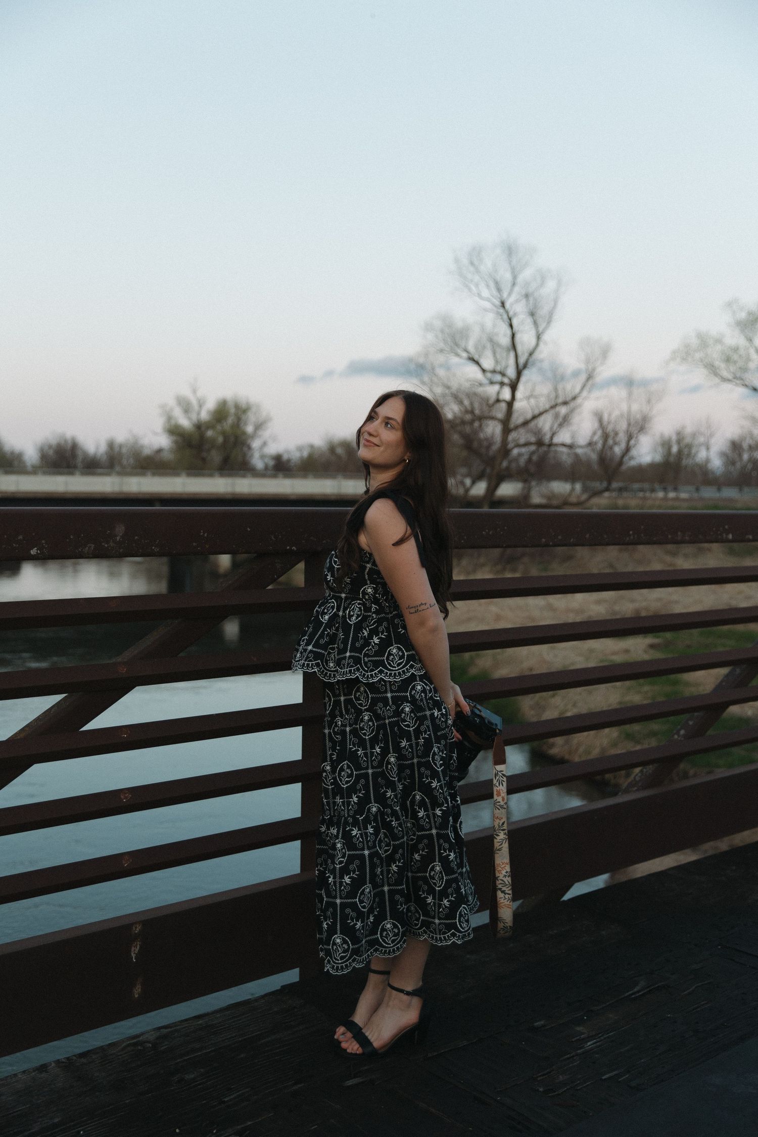 Young woman in black floral dress posing on a wooden bridge at dusk.