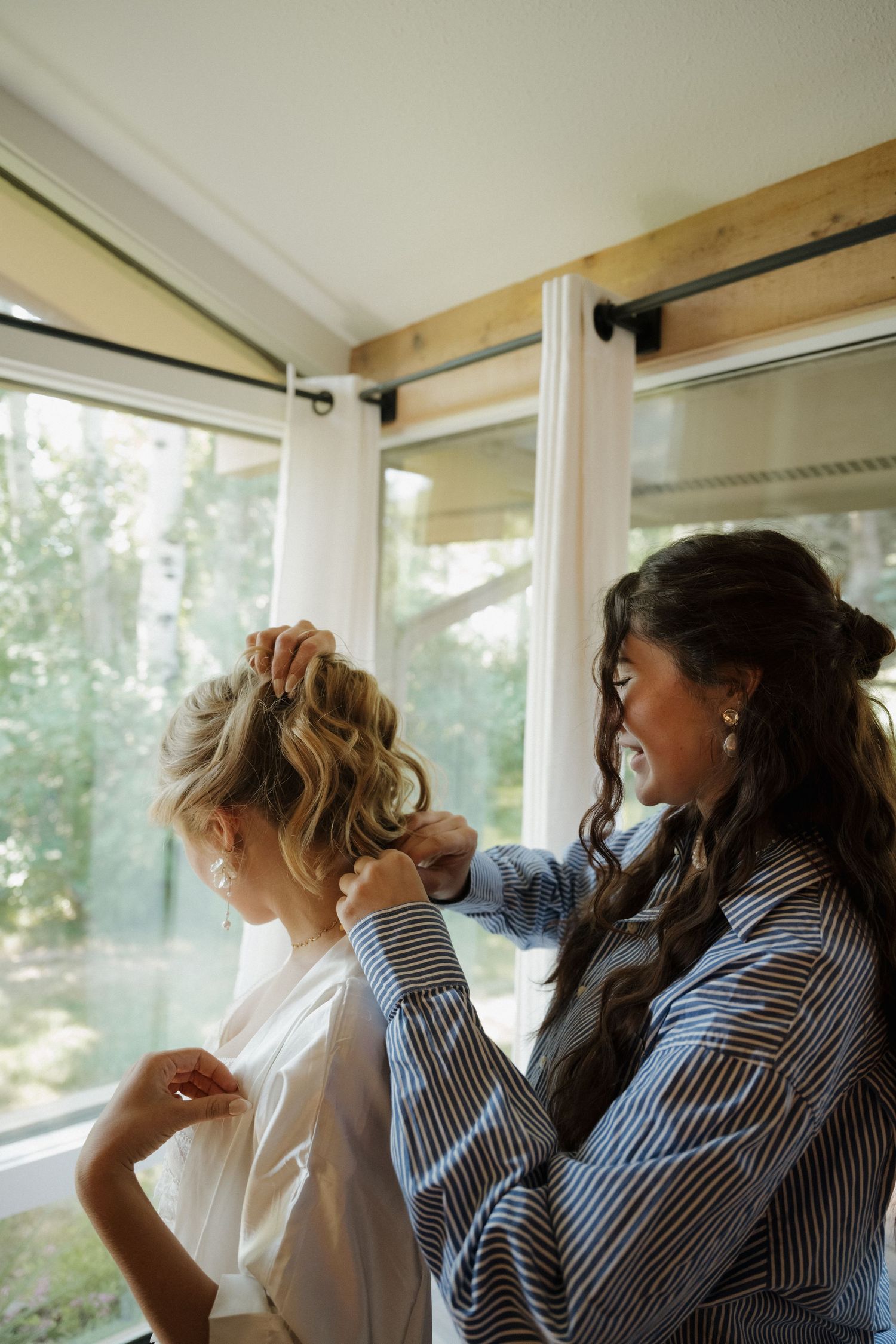 Hairstylist pinning up blonde brides elegant updo hairstyle by bright window during wedding morning prep.