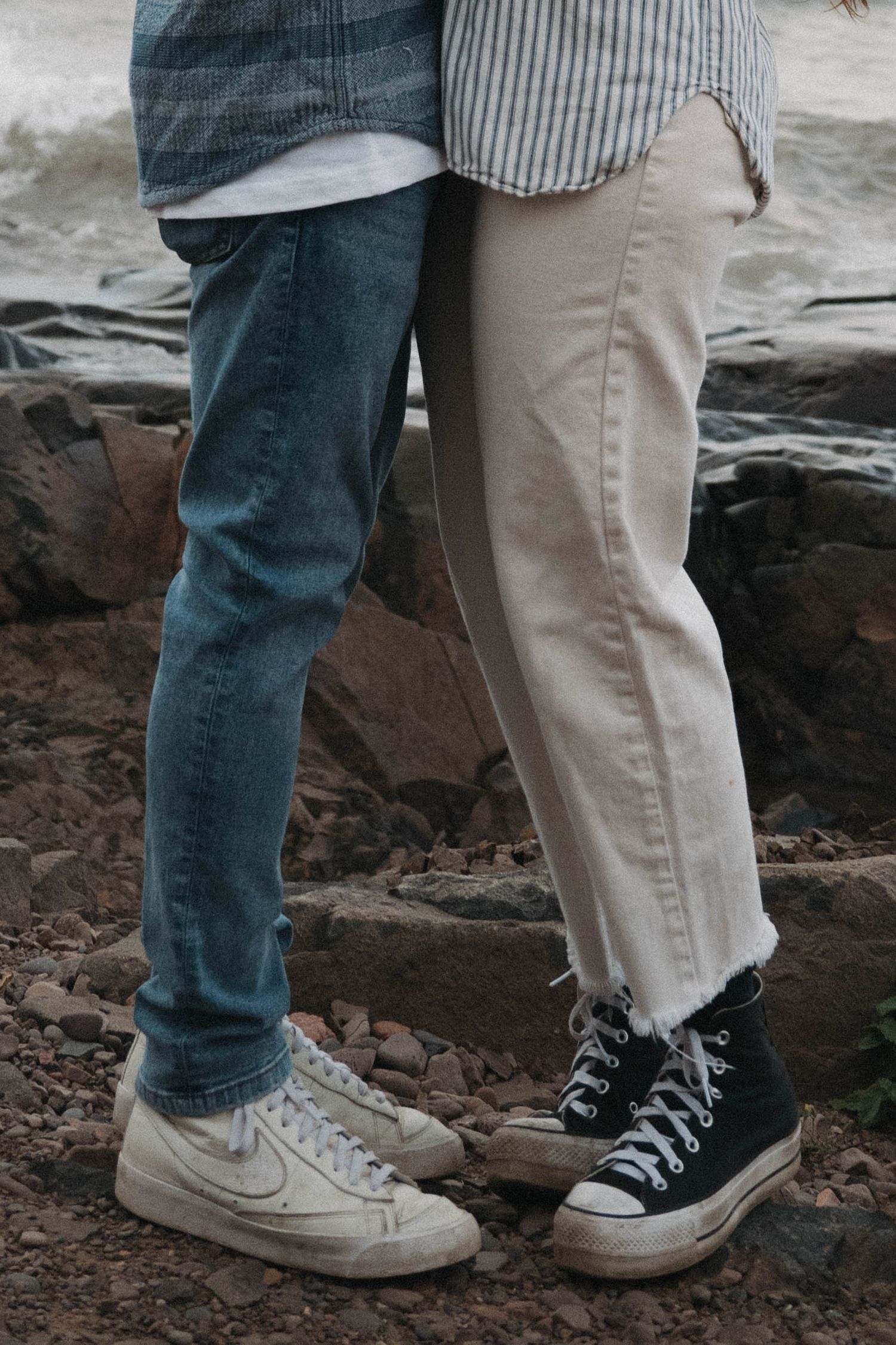 Close-up of couple's feet standing on rocky beach, wearing Nike and Converse sneakers with jeans.