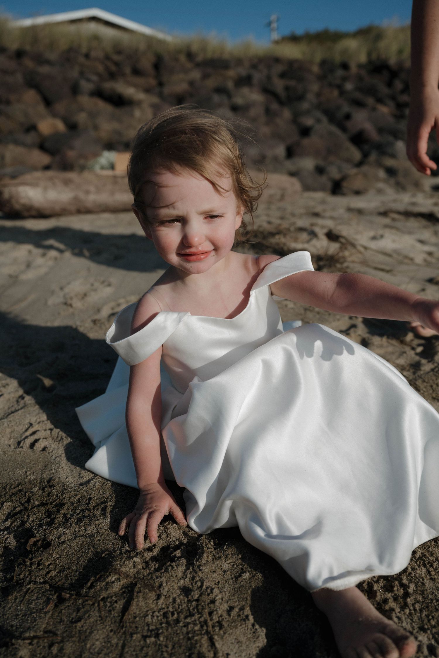 An adorable toddler in a white dress sits on sandy beach rocks smiling at the camera.