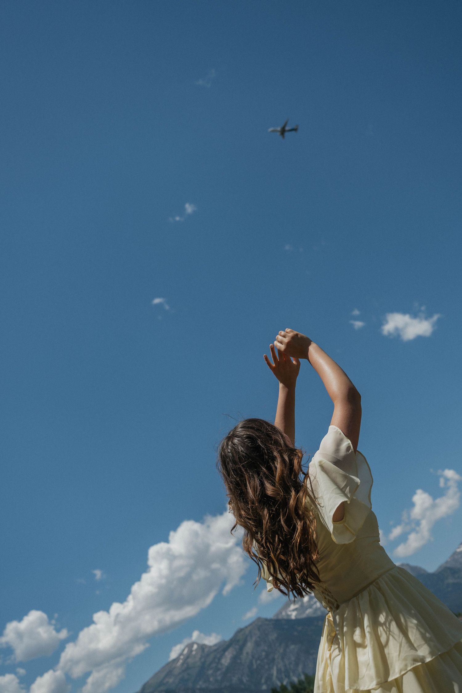 Woman in yellow dress reaching arms toward blue sky watching airplane fly over mountain landscape.