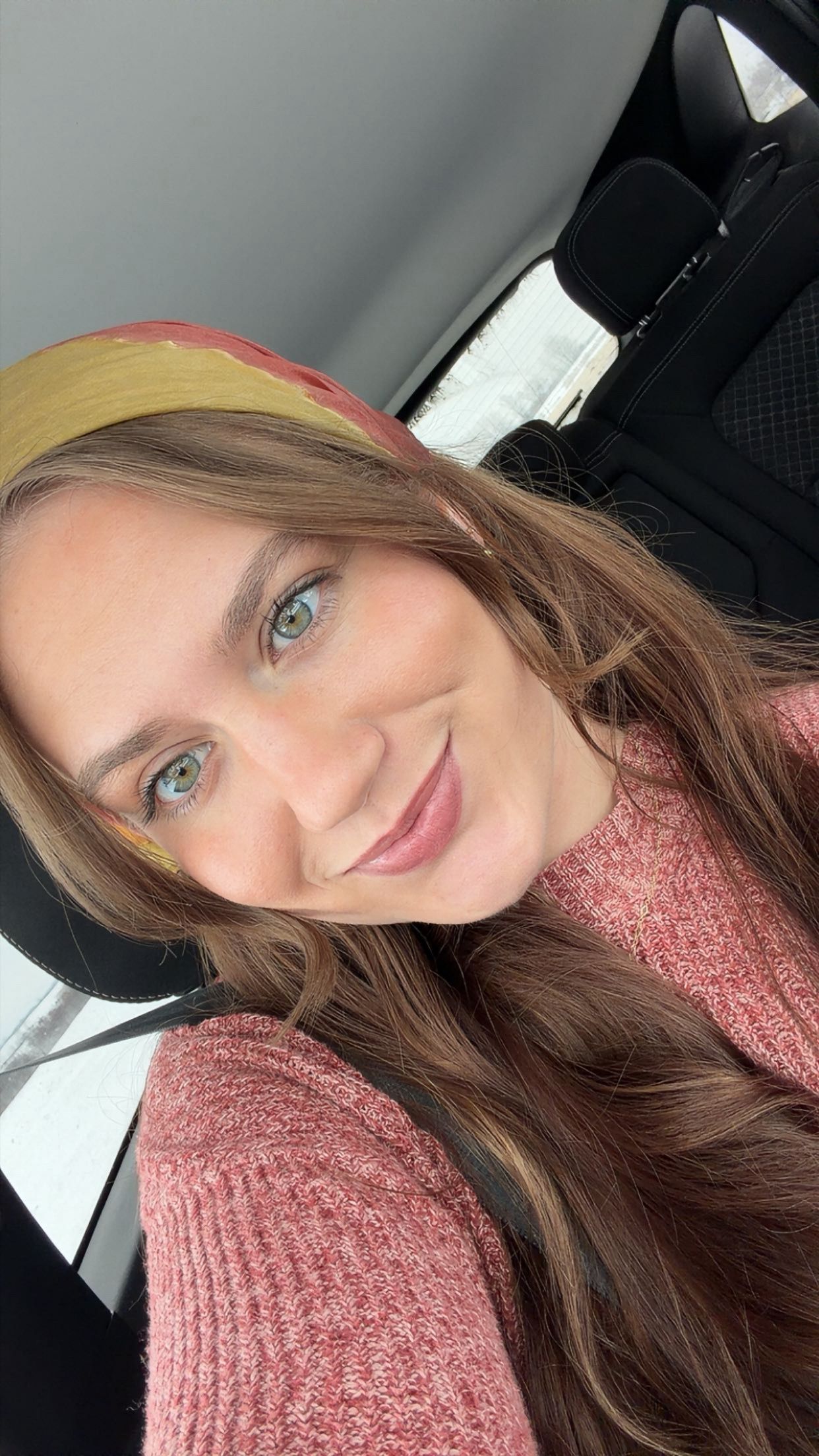 Young woman with blue eyes and brown hair taking selfie in car wearing pink sweater.