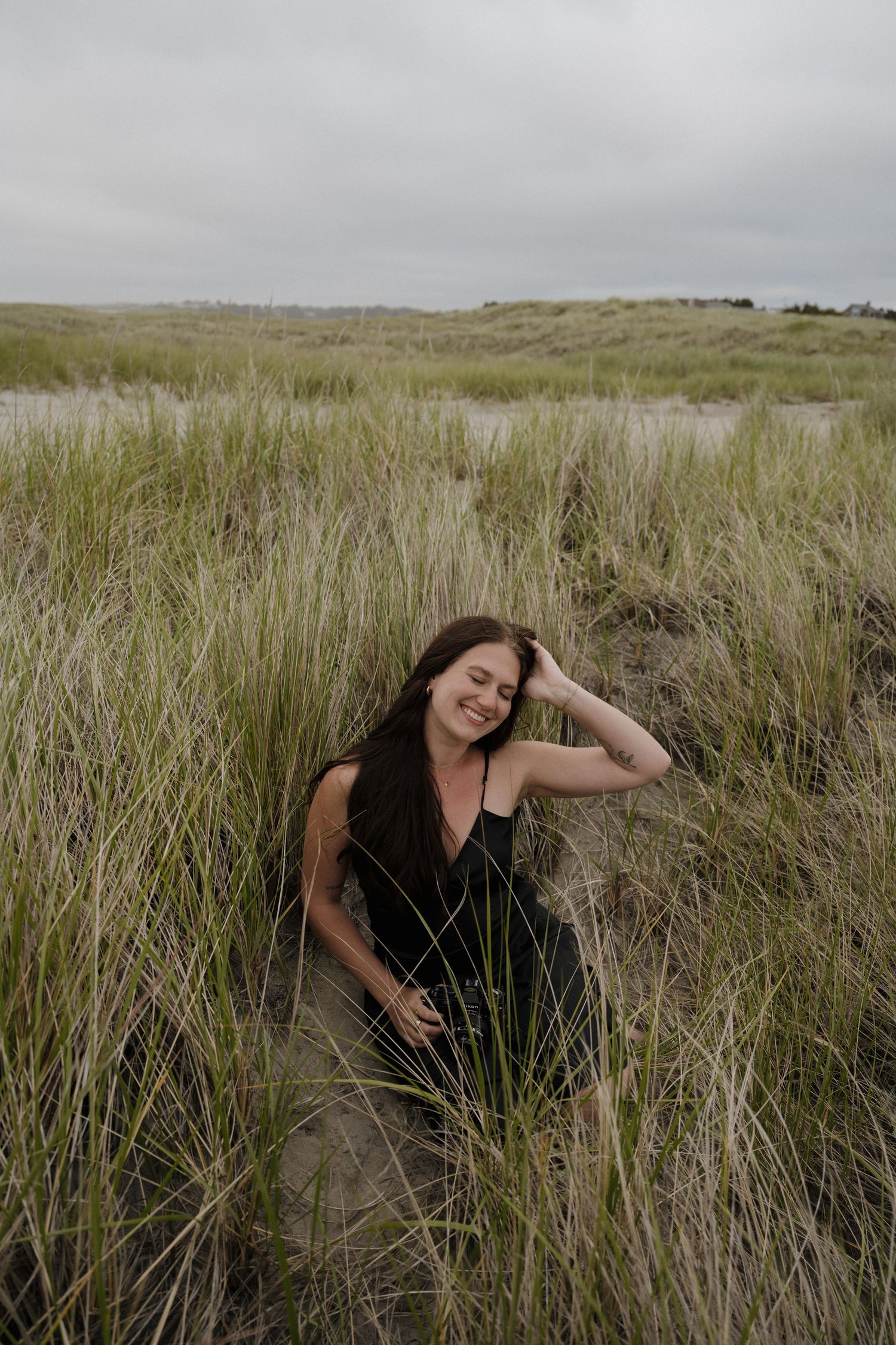 Young woman in black dress sitting relaxed among tall coastal grass on cloudy day.