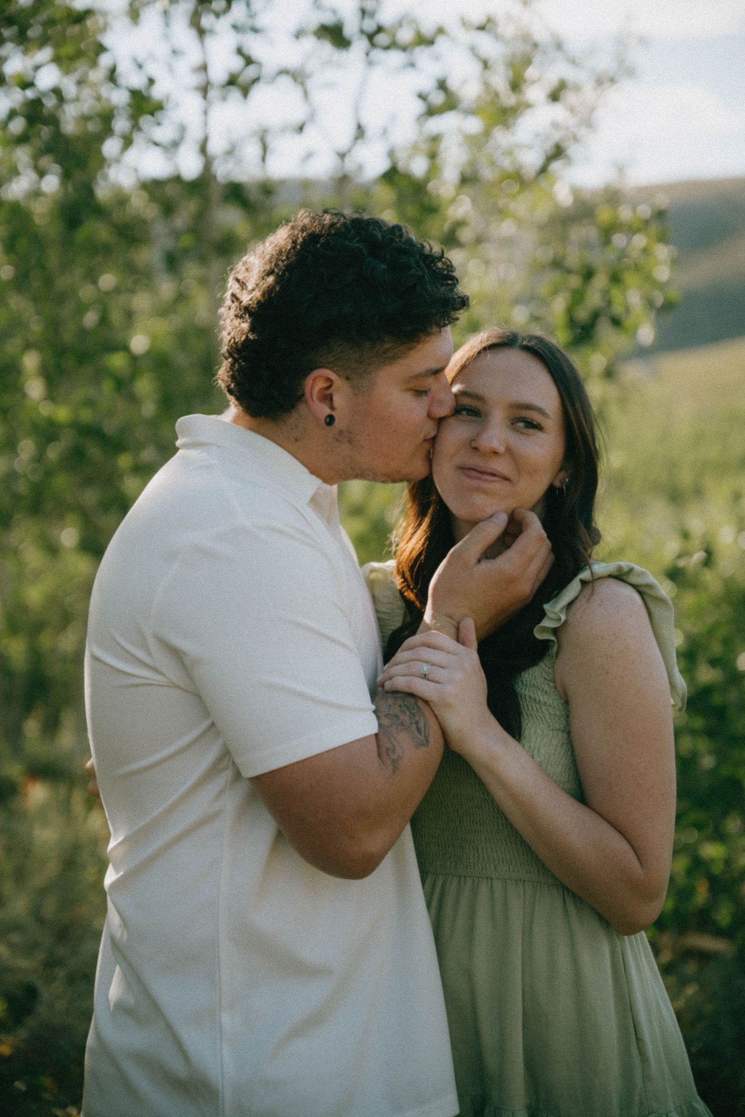 A man kisses a woman on the cheek while holding her face tenderly in a lush green outdoor setting.