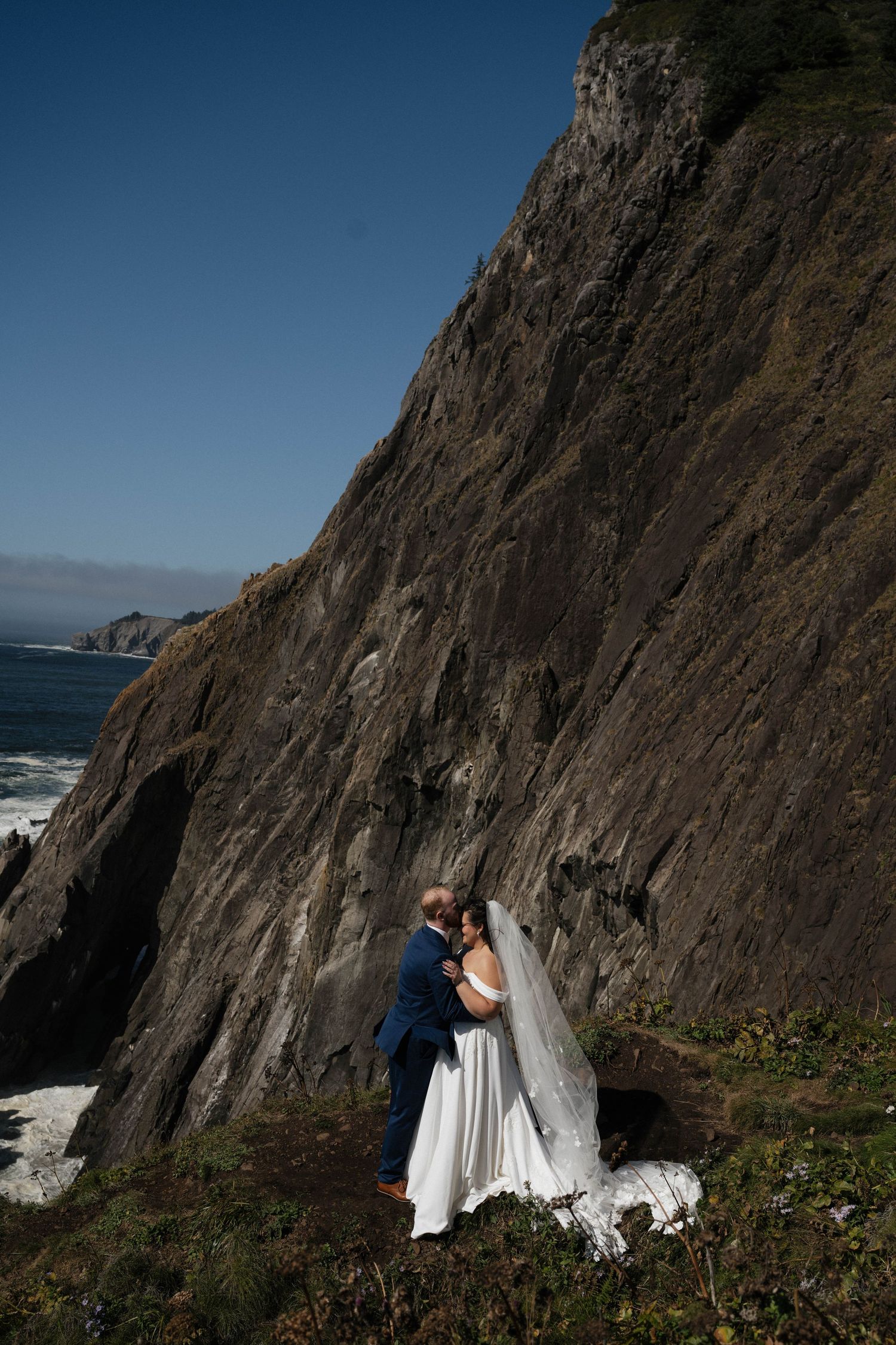 Bride and groom embrace romantically against dramatic rocky coastal cliffs with ocean waves below on their wedding day.