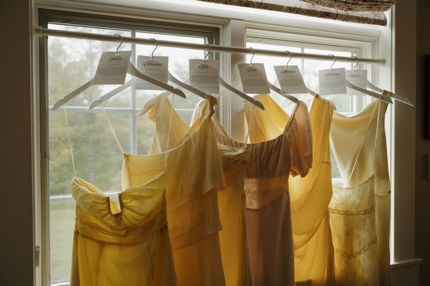 Yellow and gold bridesmaid dresses hanging on hangers in front of a bright window.
