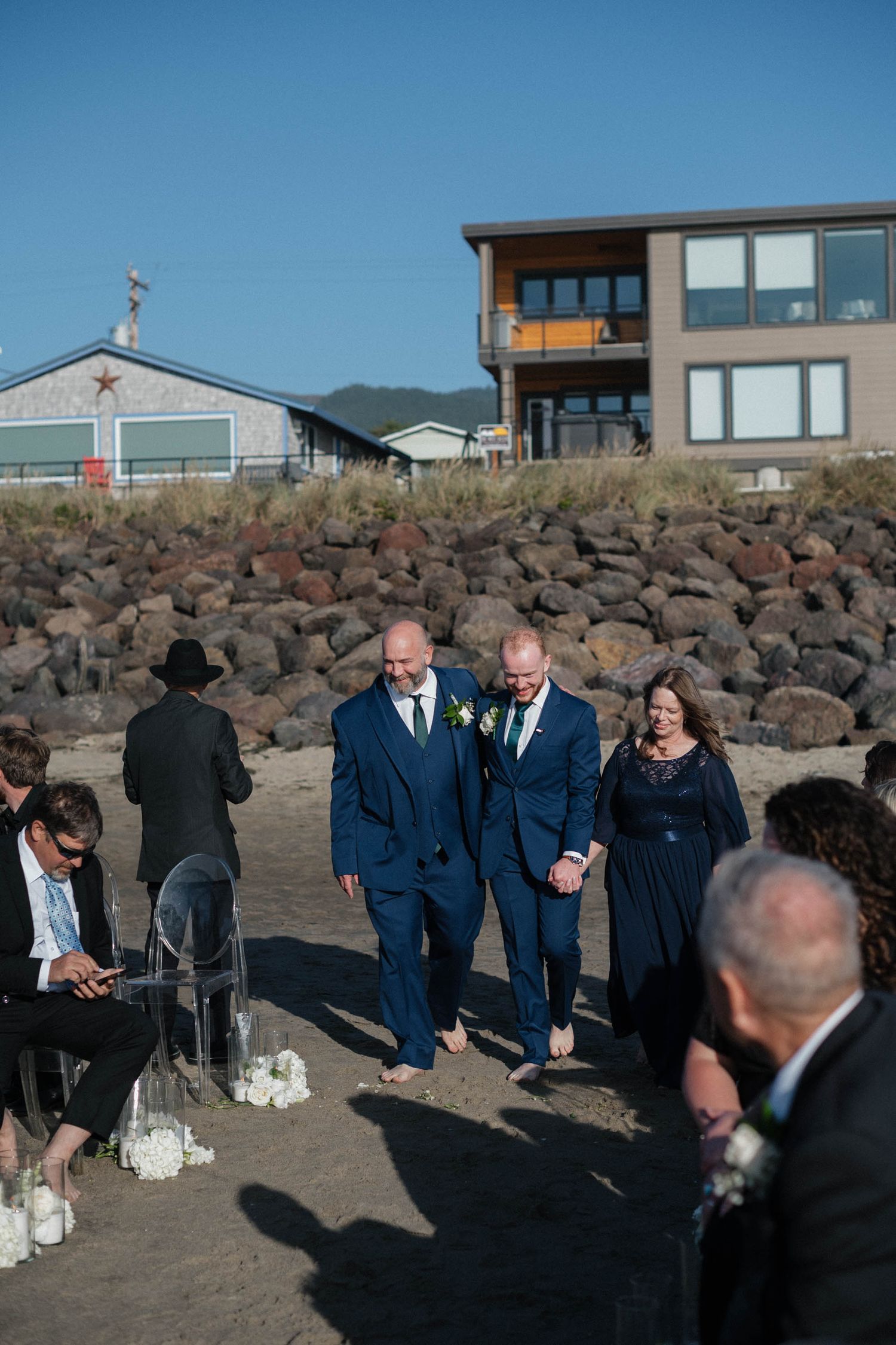 Two grooms in navy suits walk down the aisle at an outdoor beach wedding ceremony.