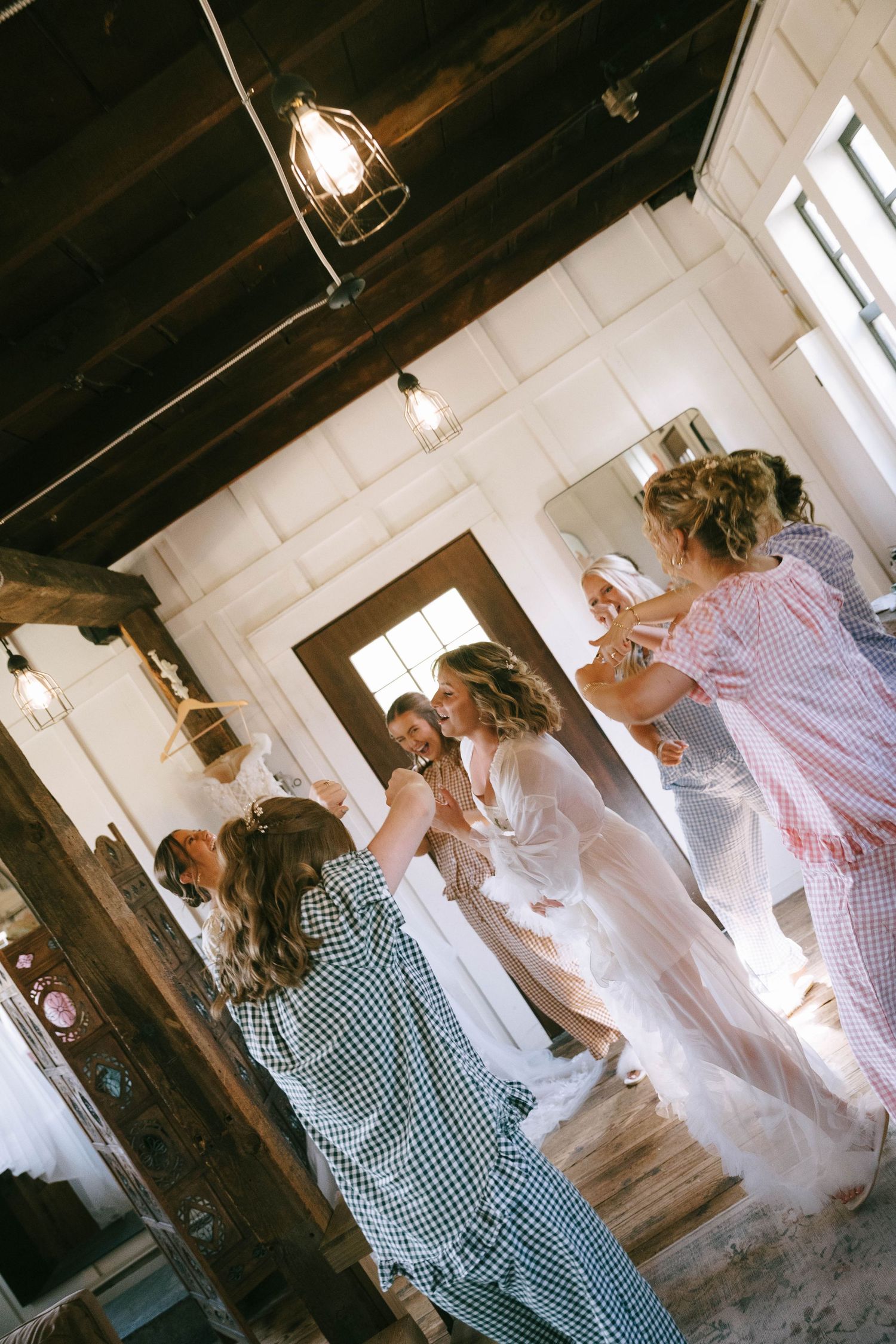 Bridesmaids helping bride into wedding dress in rustic white room with wood ceiling and pendant lights.