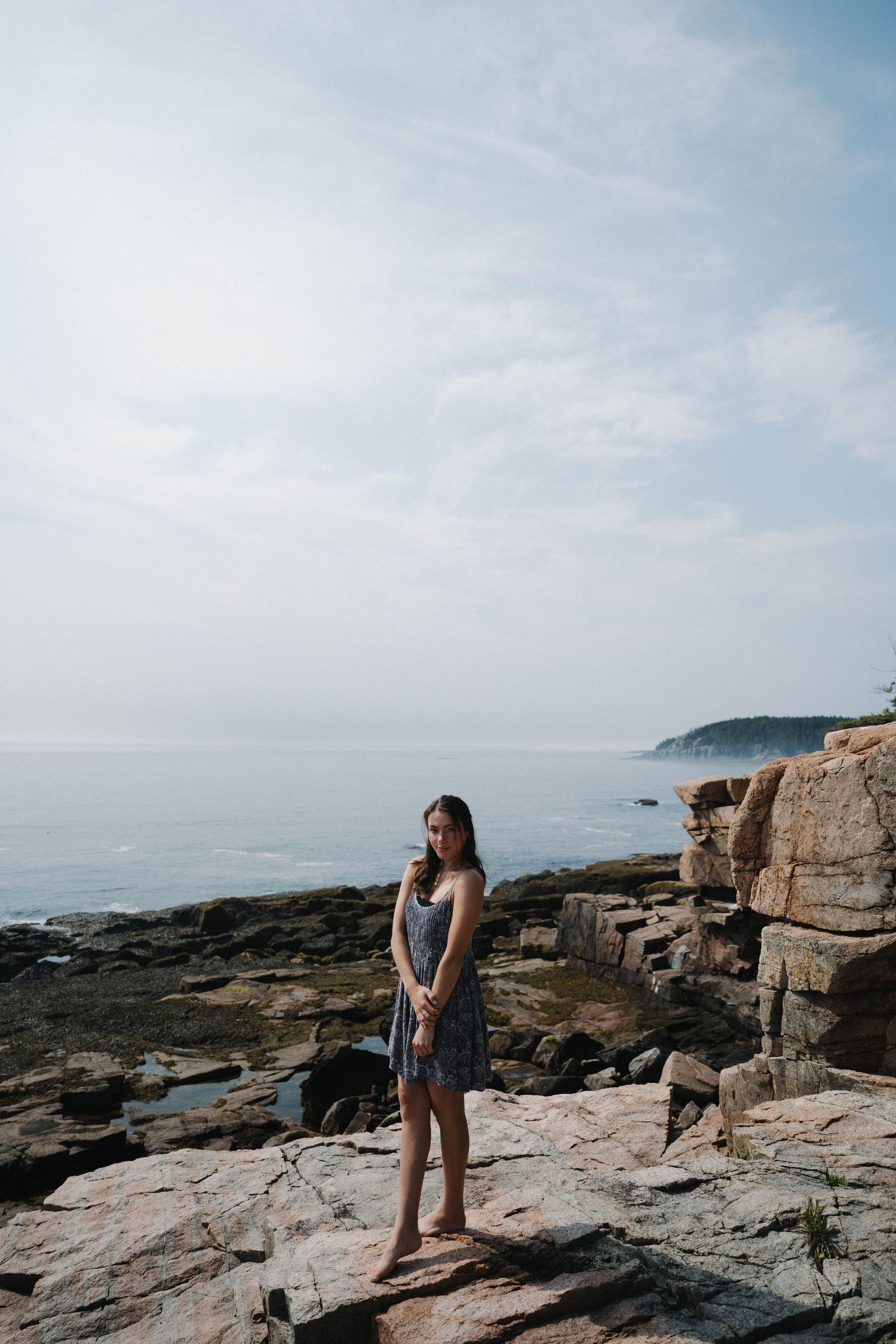 Young woman in floral dress standing on rocky coastal cliffs with ocean and misty headlands in background.
