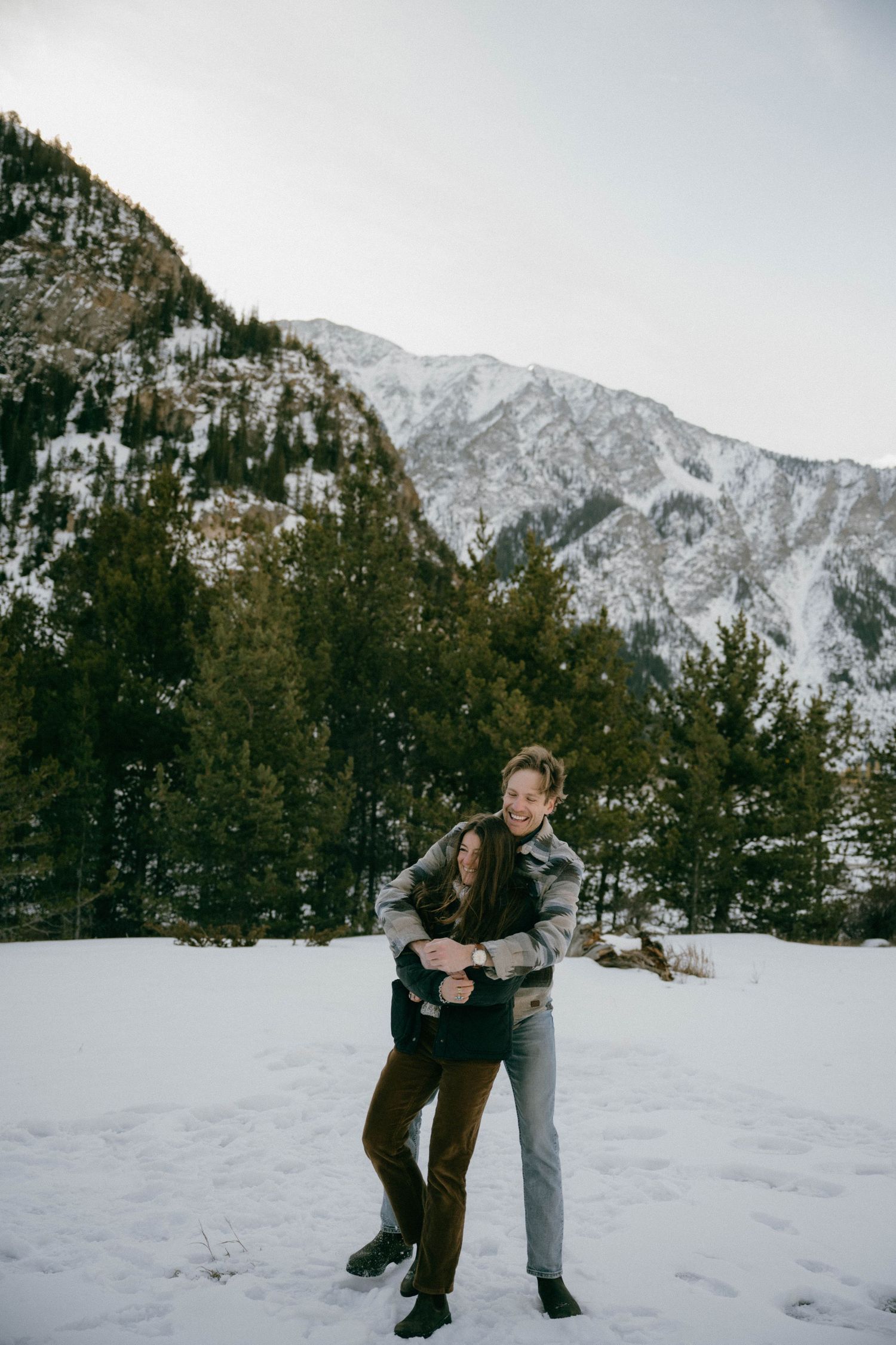 Couple laughs and embraces playfully in snowy mountain landscape surrounded by evergreen trees.
