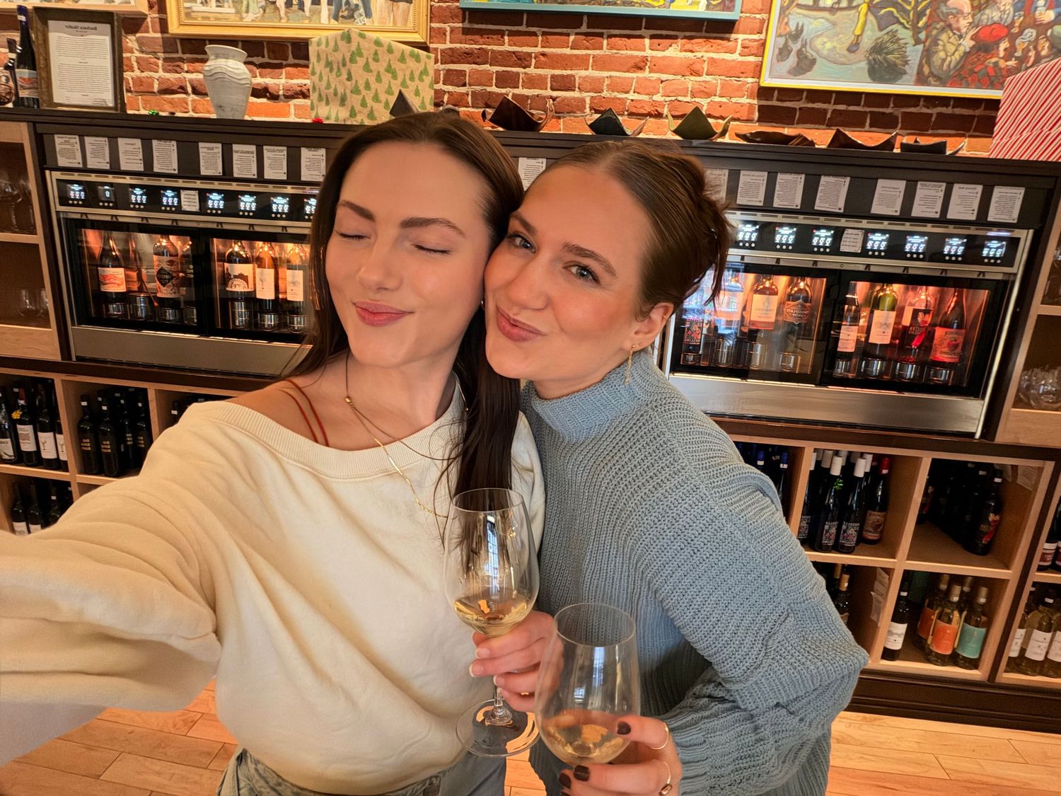 Two women smiling and holding white wine glasses at a wine bar with bottles displayed on shelves behind them.