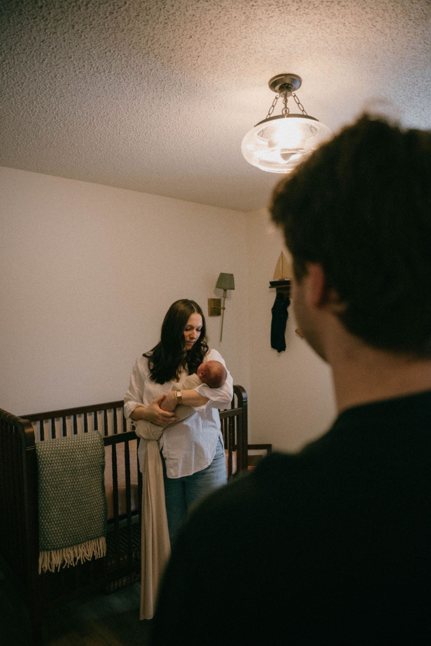 Mother holds newborn baby in nursery while father watches lovingly nearby.
