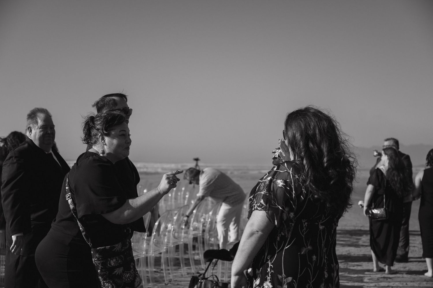 Black and white photo of wedding guests mingling near clear chairs at a beach ceremony.