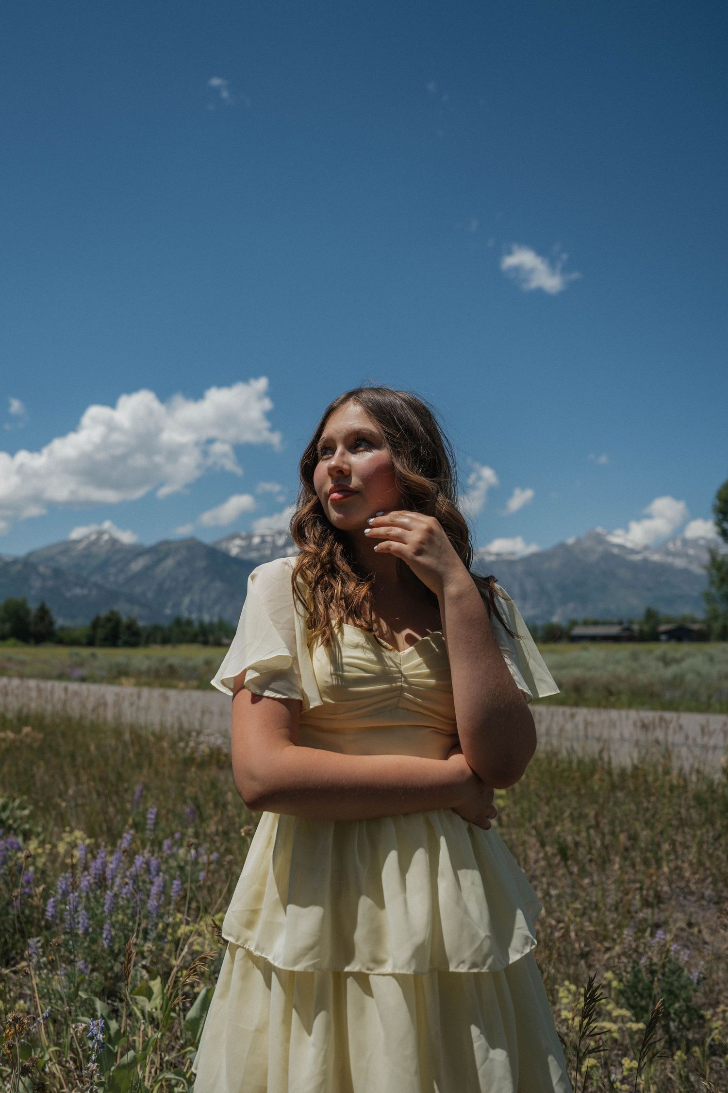 Young woman in cream tiered dress posing thoughtfully in wildflower meadow with mountain skyline.