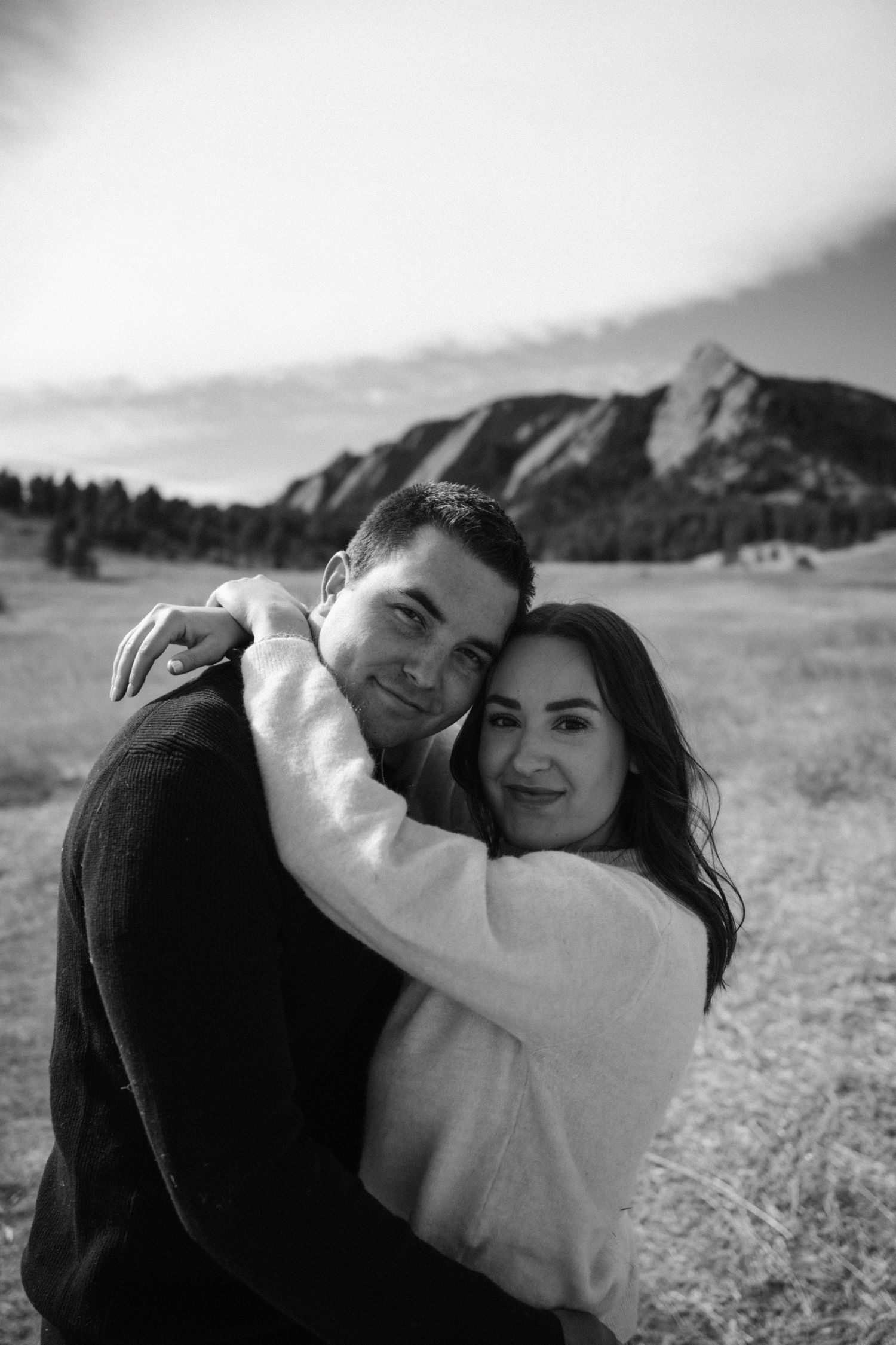 Black and white portrait of smiling couple embracing warmly with Colorado Flatirons in background.
