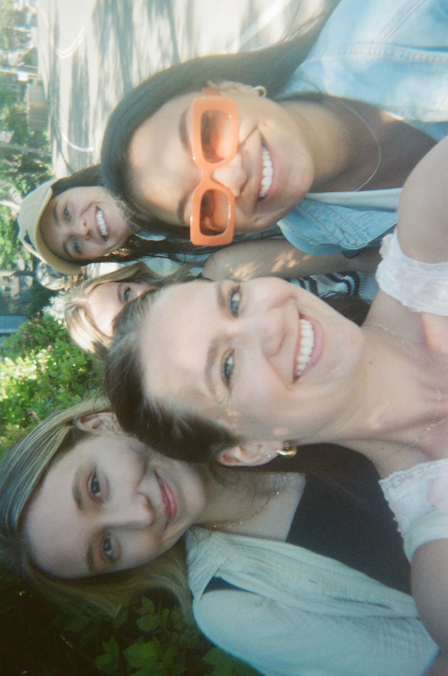 Four smiling young women taking a fun outdoor selfie, one wearing orange sunglasses on a sunny day.