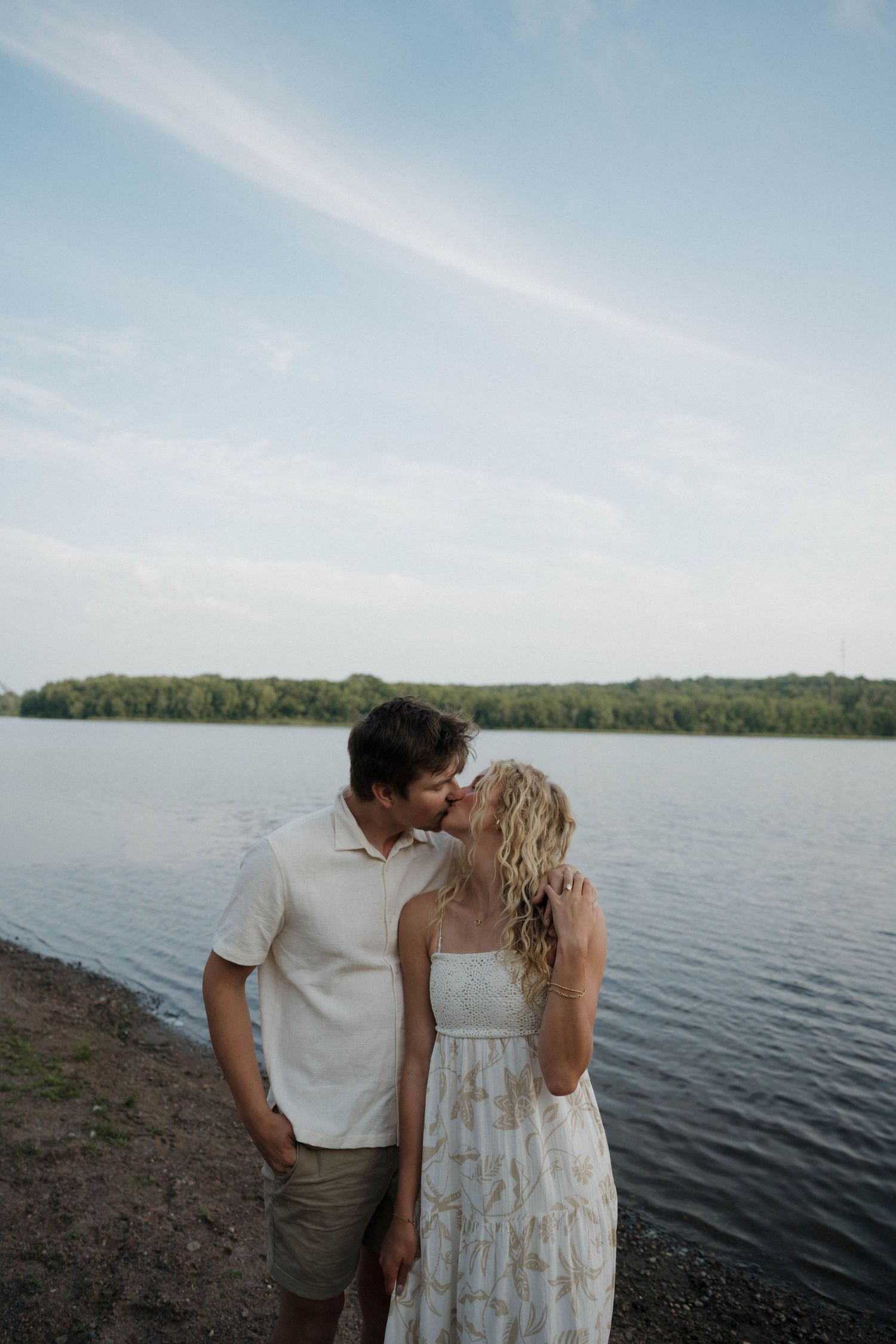 Couple sharing a kiss by a calm lake at sunset with treeline in background.