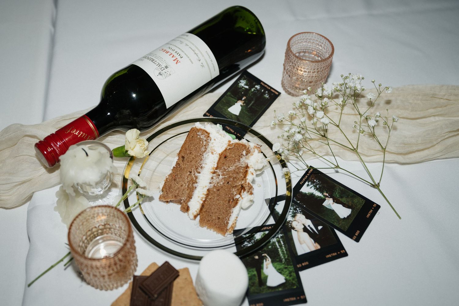 Wedding table with wine bottle, cake slice, photos, and floral decor.