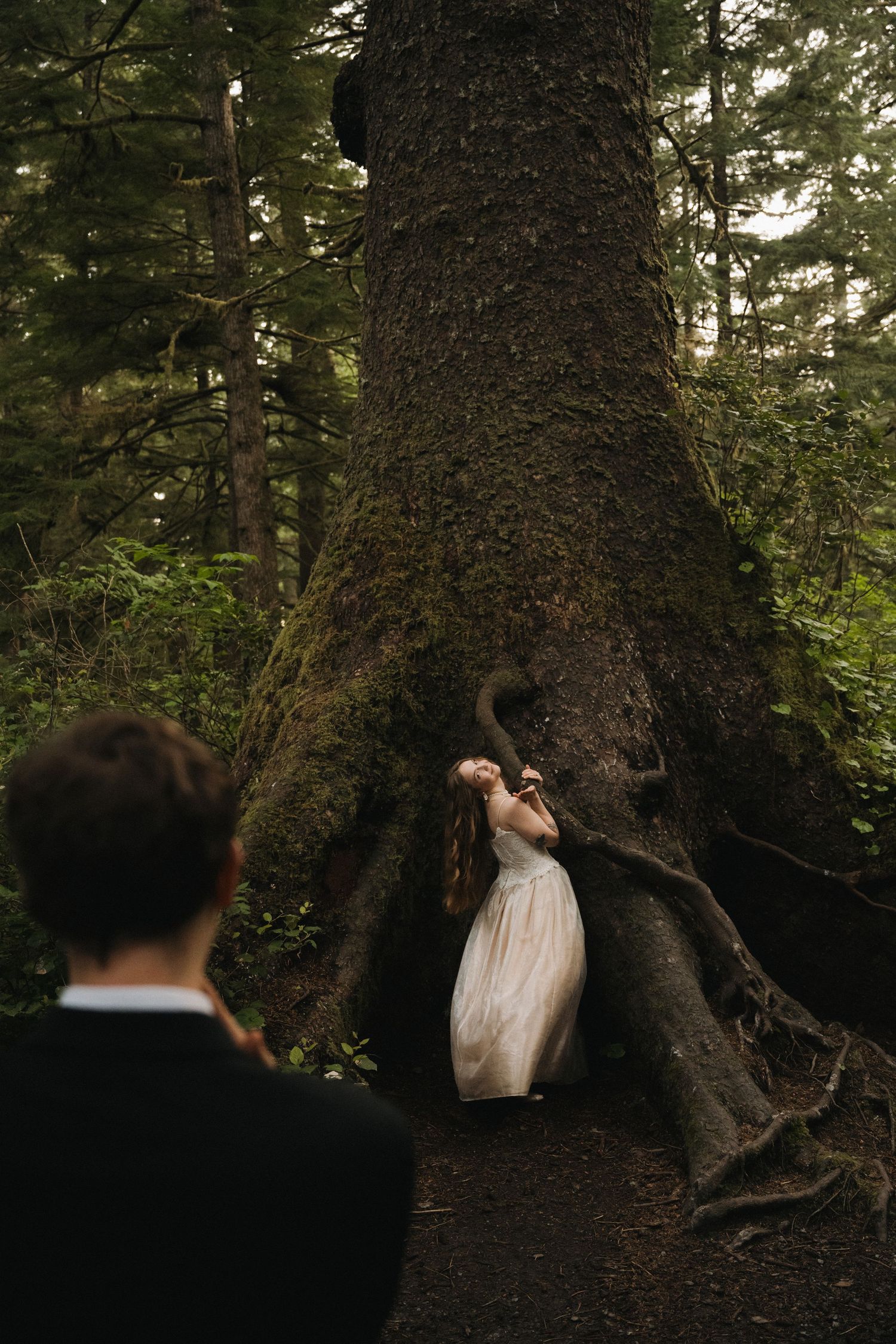 Bride in white gown leans against massive moss-covered tree trunk in lush forest setting.