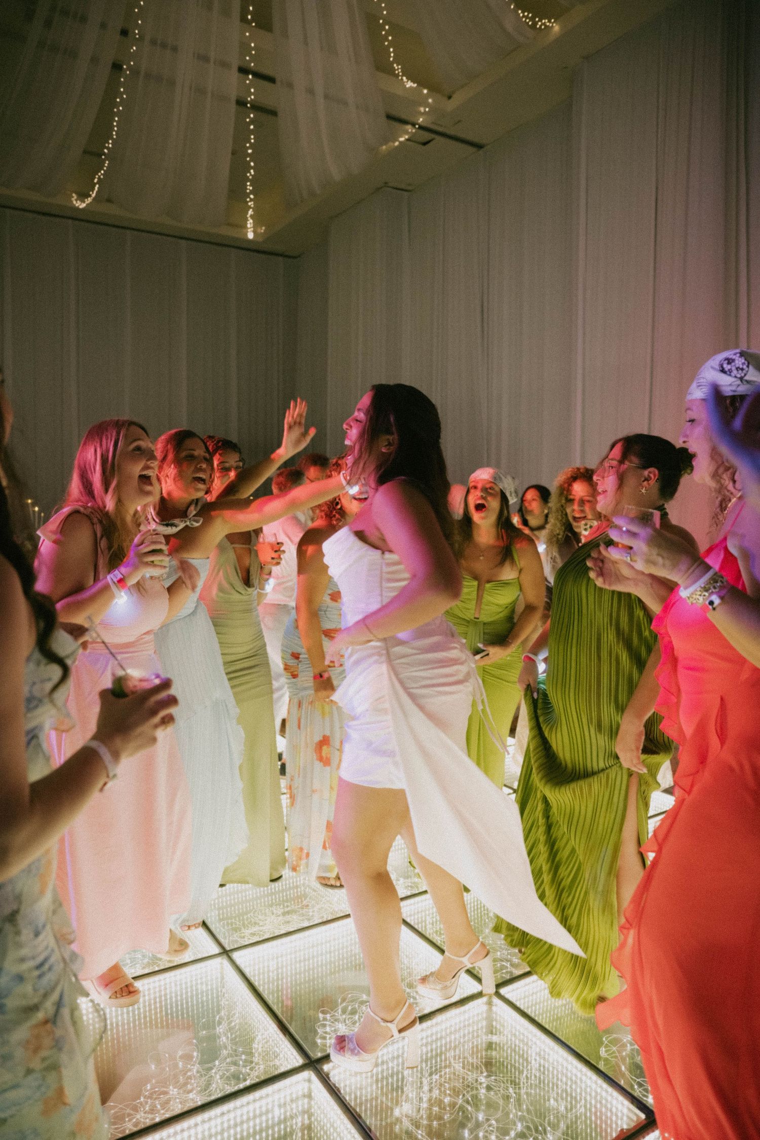 Bride dancing joyfully with bridesmaids in colorful dresses on illuminated dance floor at wedding reception.