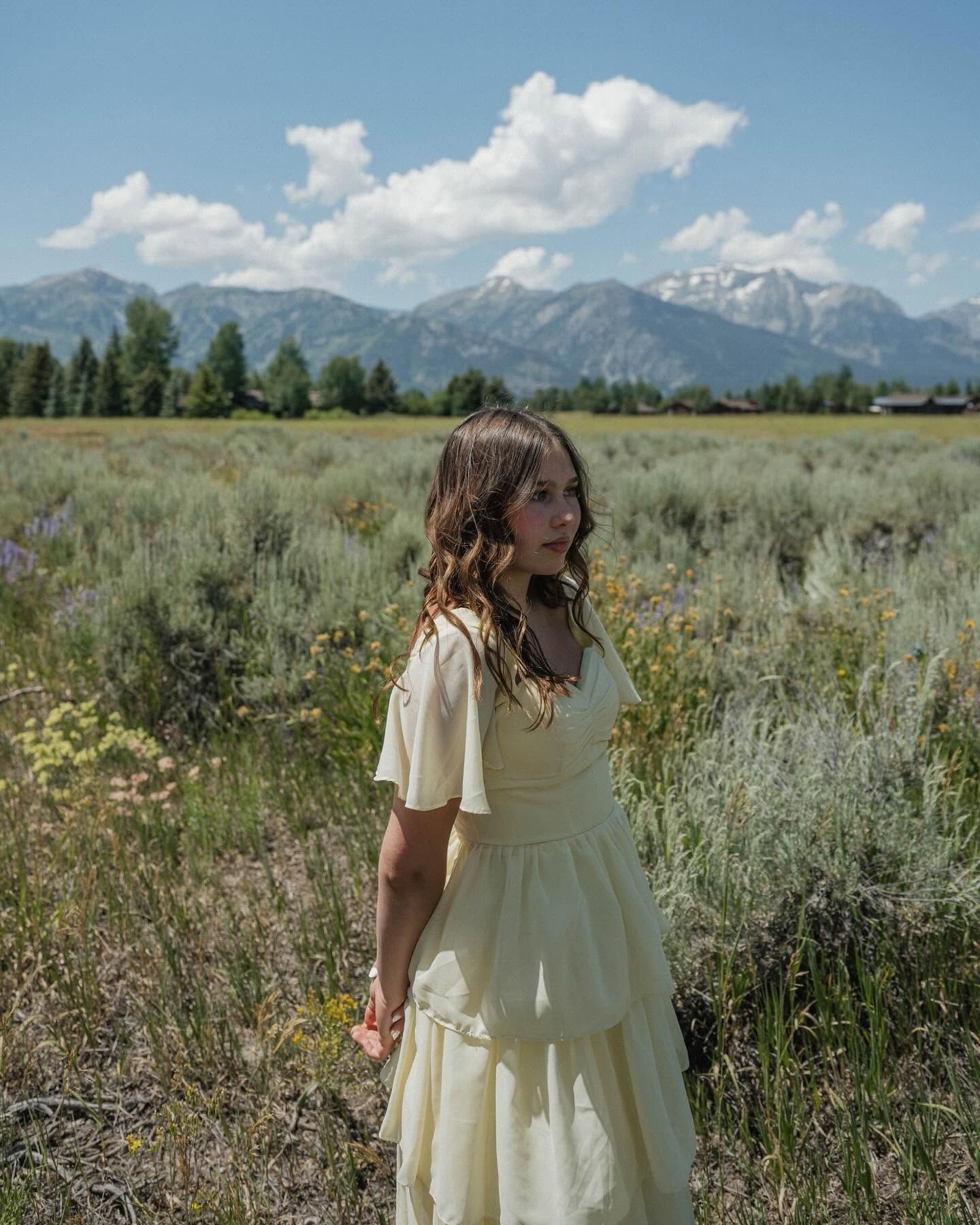 Young woman in a cream dress stands in a wildflower field with snow-capped mountains in the background.