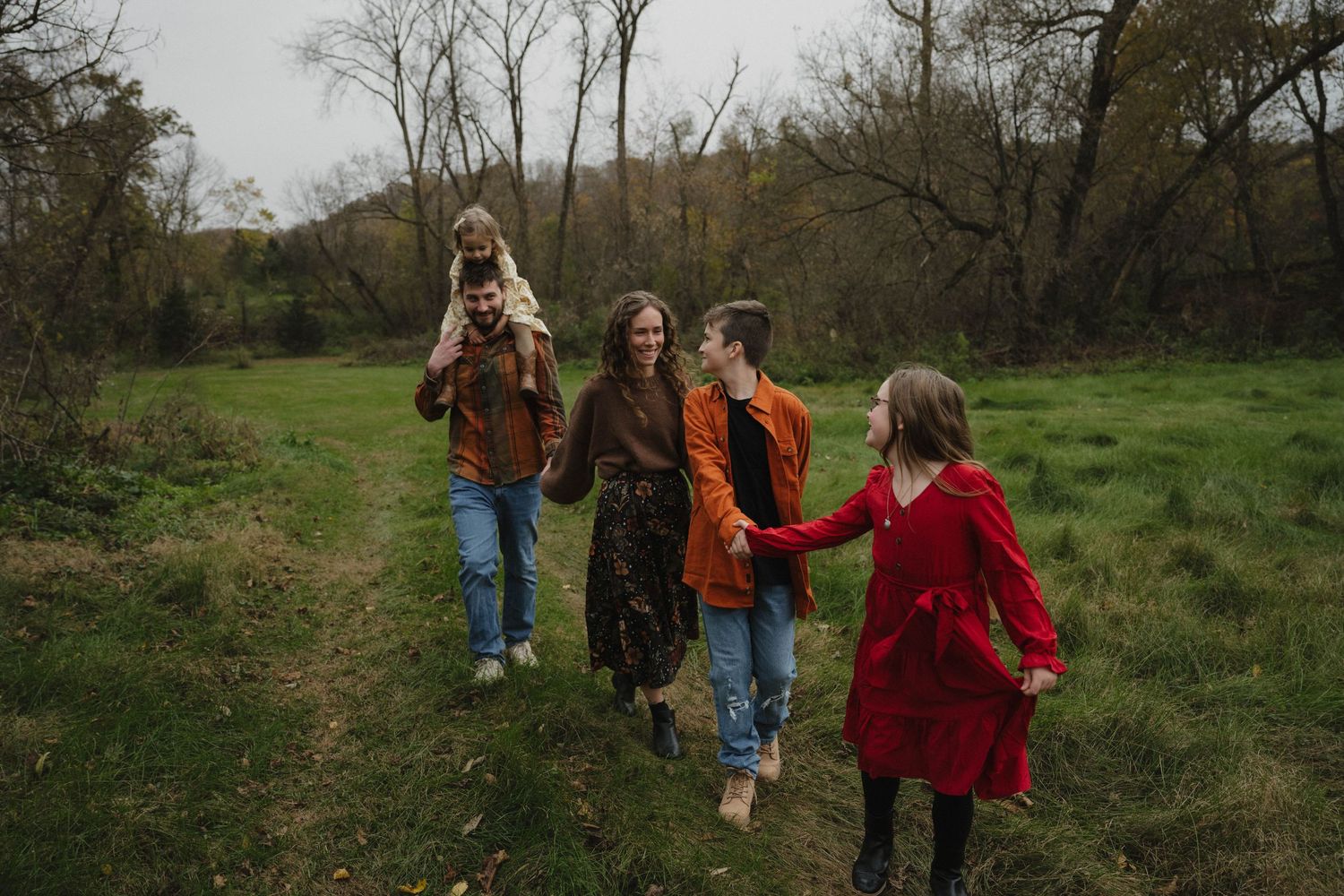 Family of four walking through a lush green meadow path surrounded by autumn trees in warm fall clothing.