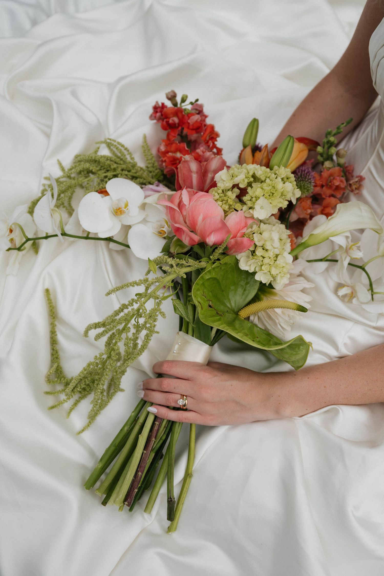 Bride holding a colorful wedding bouquet with pink, red, and white flowers on white satin fabric, wearing an engagement ring.