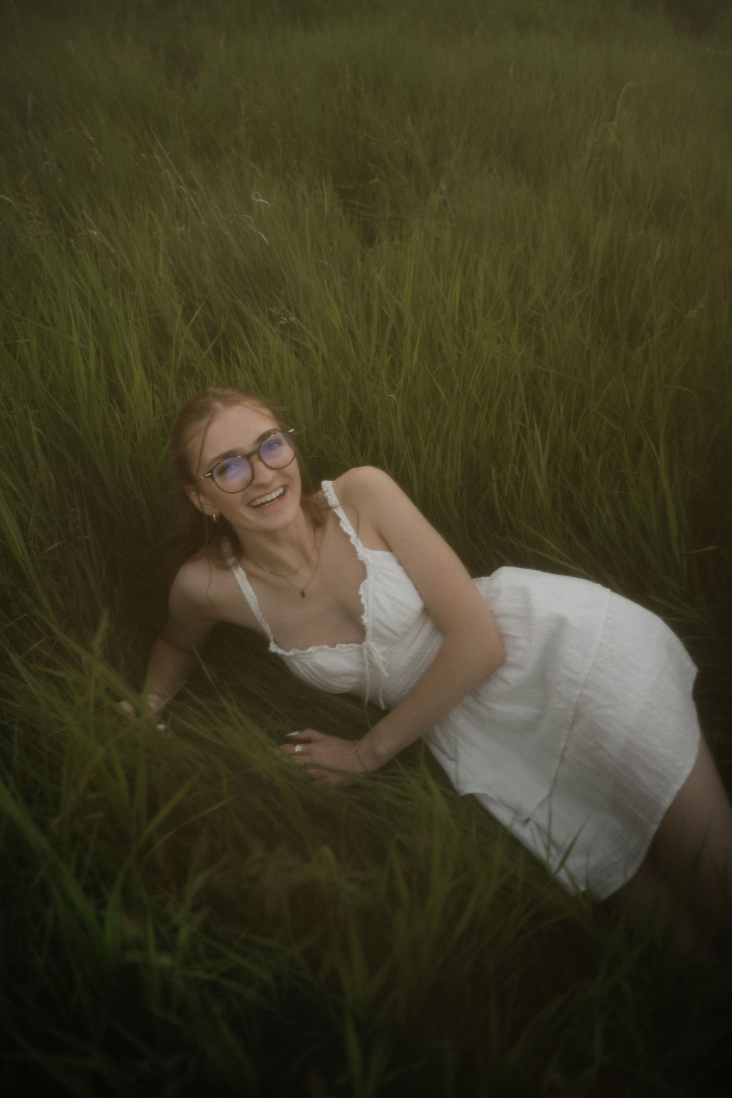 Laughing young woman with glasses in a white dress lies in tall green grass during an outdoor photo session.