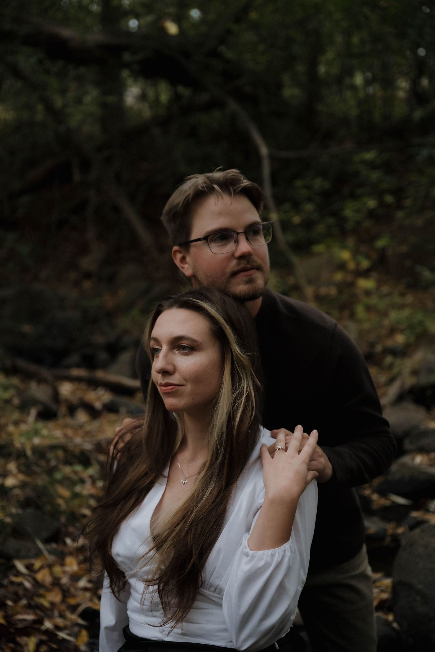 A man with glasses embraces a woman with long hair in a moody autumn forest setting.