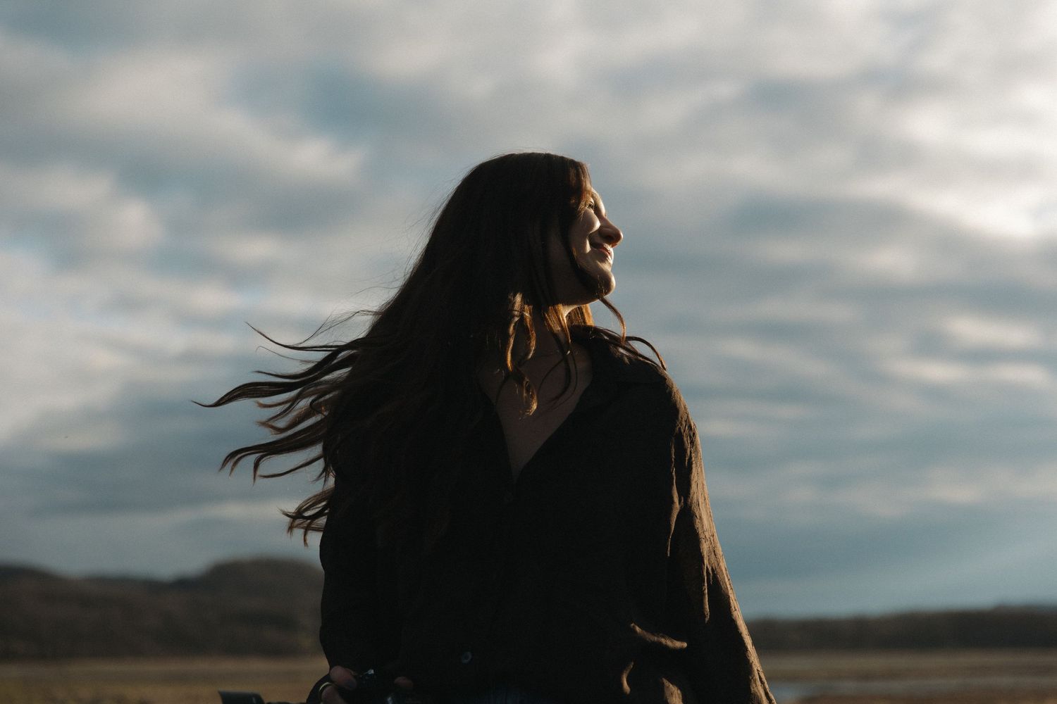 Young woman with flowing dark hair smiles serenely outdoors beneath a moody overcast sky.