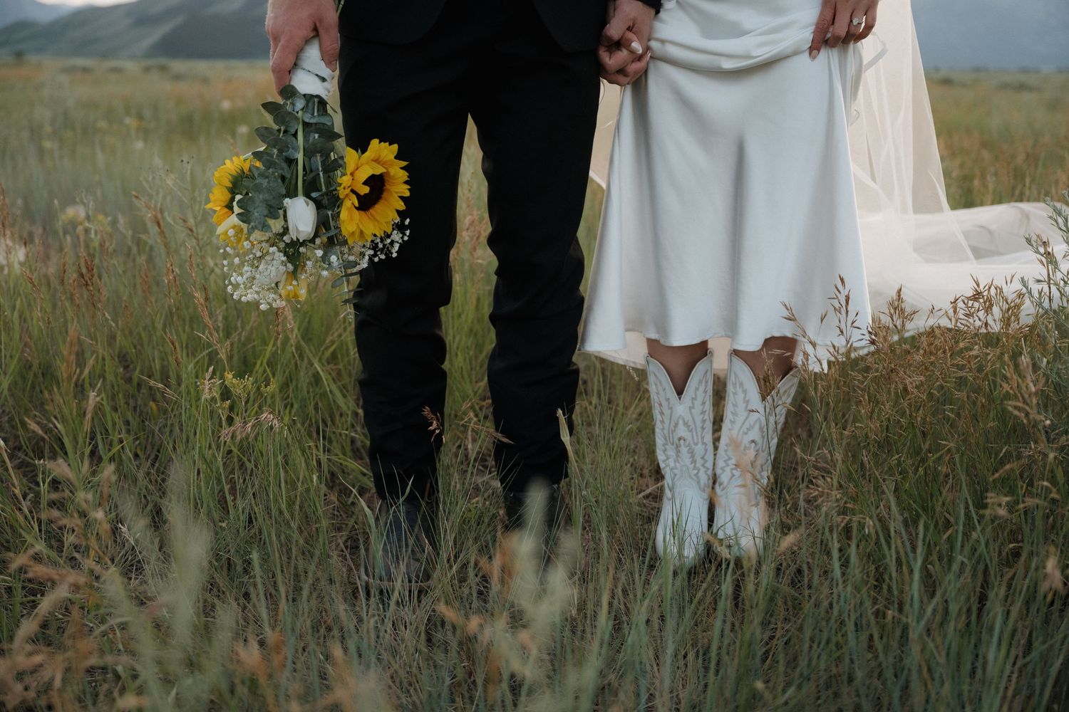 Bride in white cowboy boots and groom hold sunflower bouquet in rustic mountain field.
