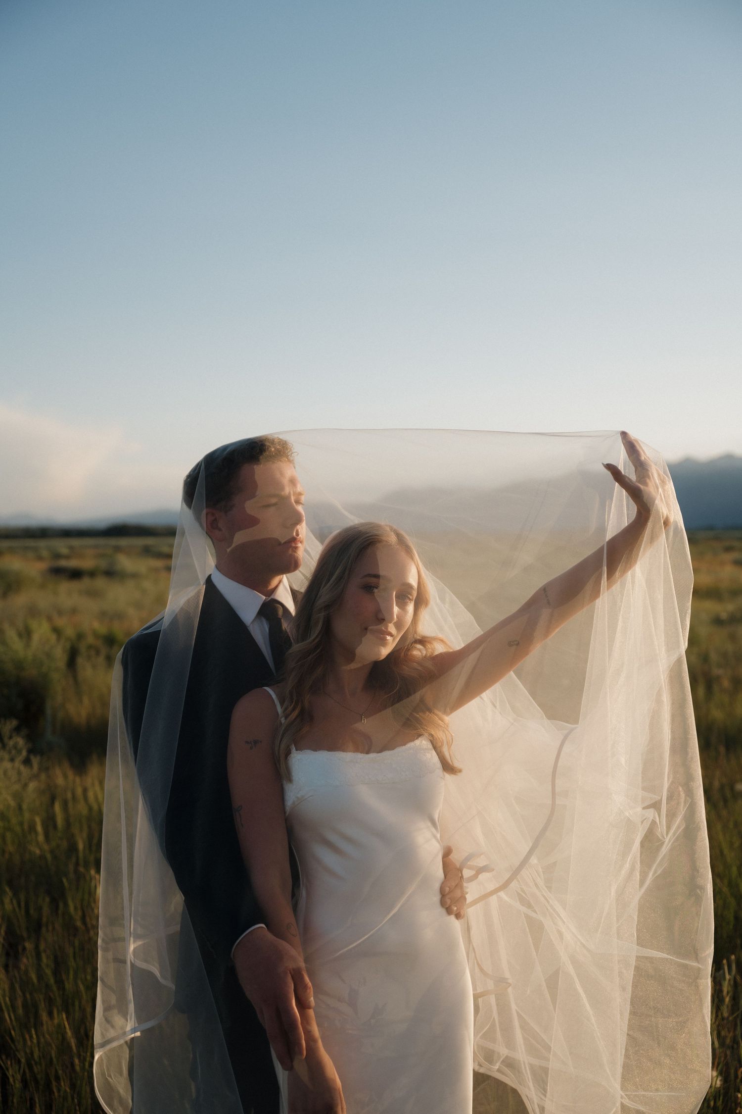 Bride lifts veil while groom stands behind her in open mountain field during golden hour.