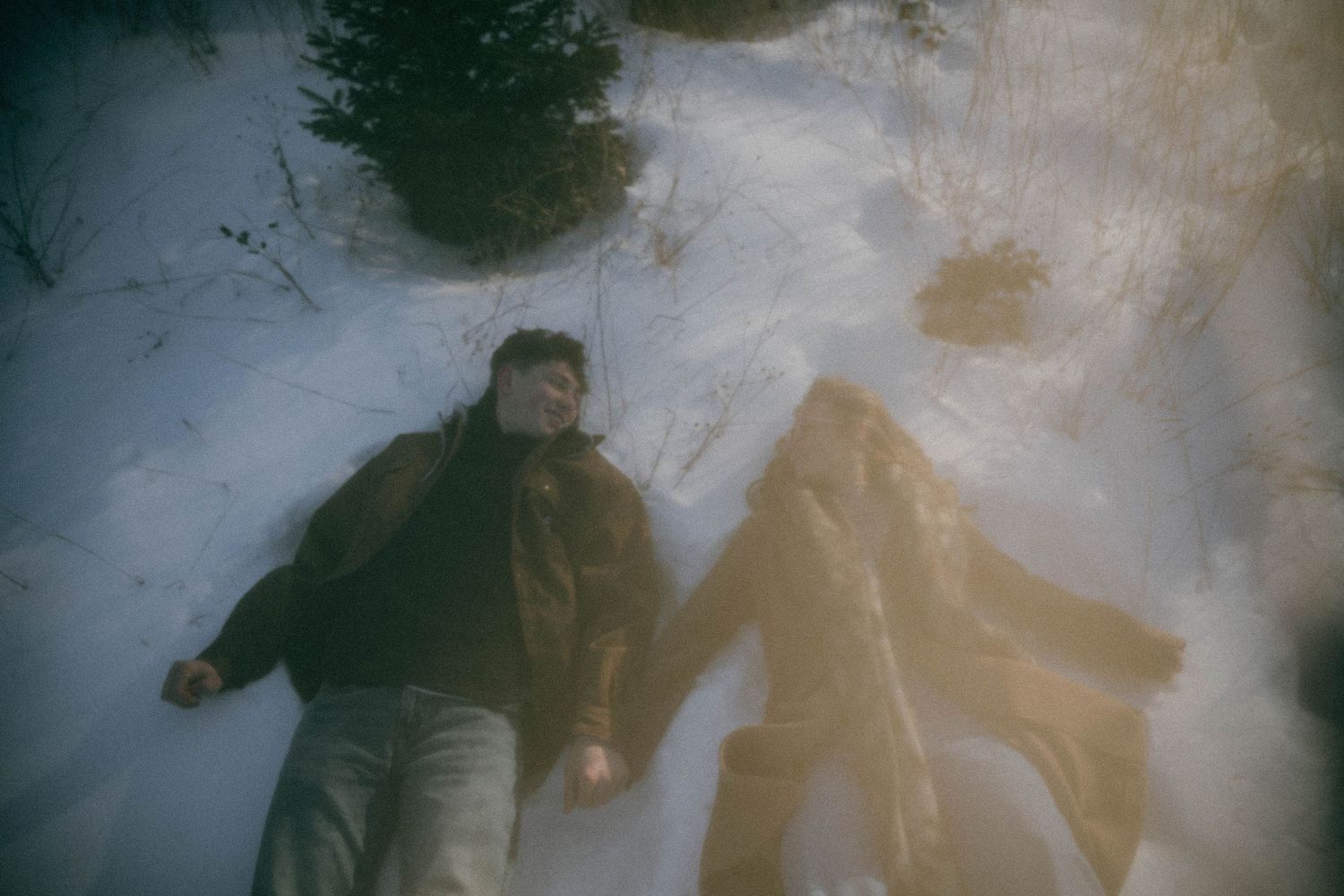 Dreamy double exposure of couple lying in snow at a winter Christmas tree farm.