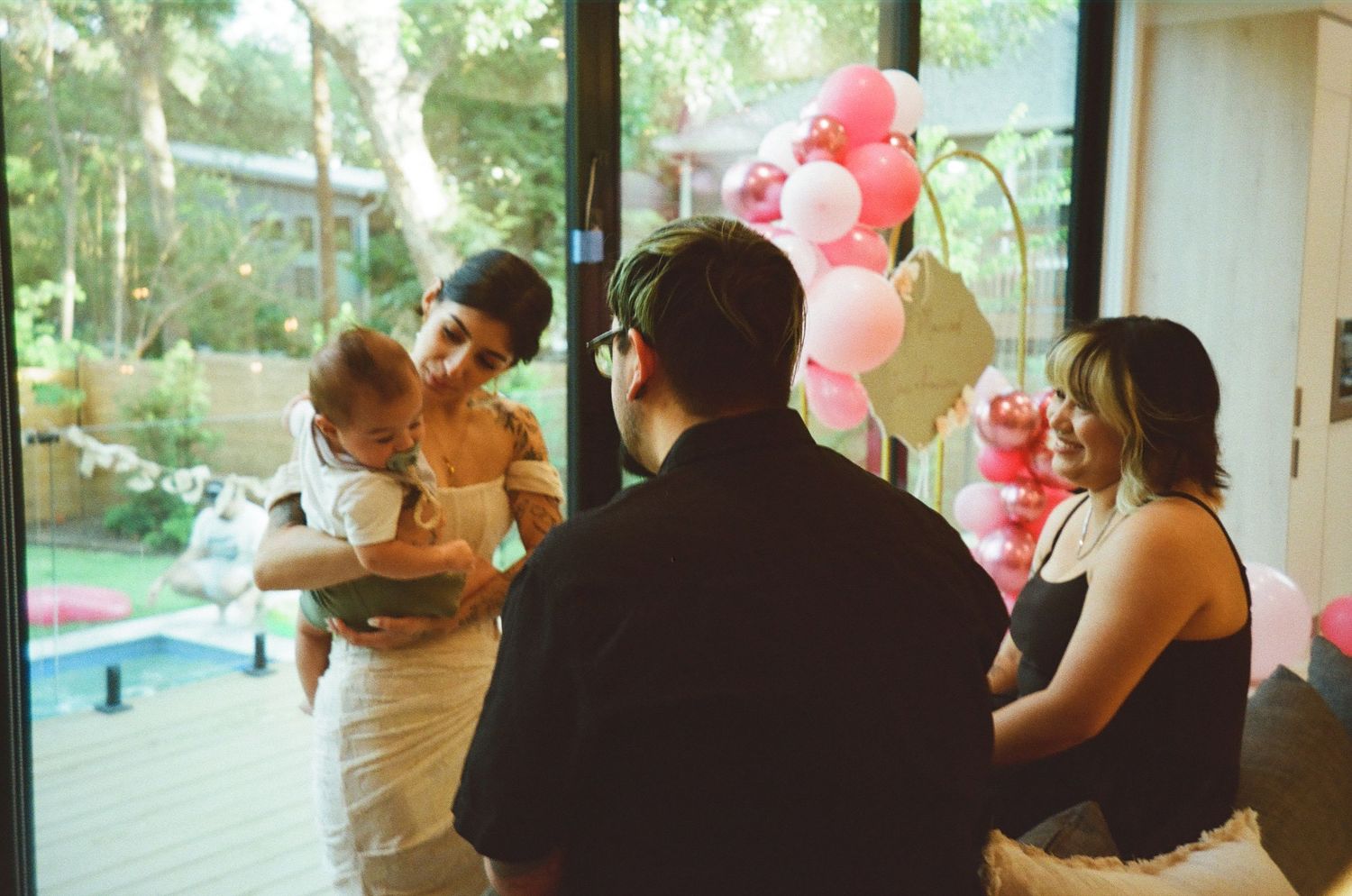Bride holding baby while guests gather near pink balloon arch during intimate wedding celebration.
