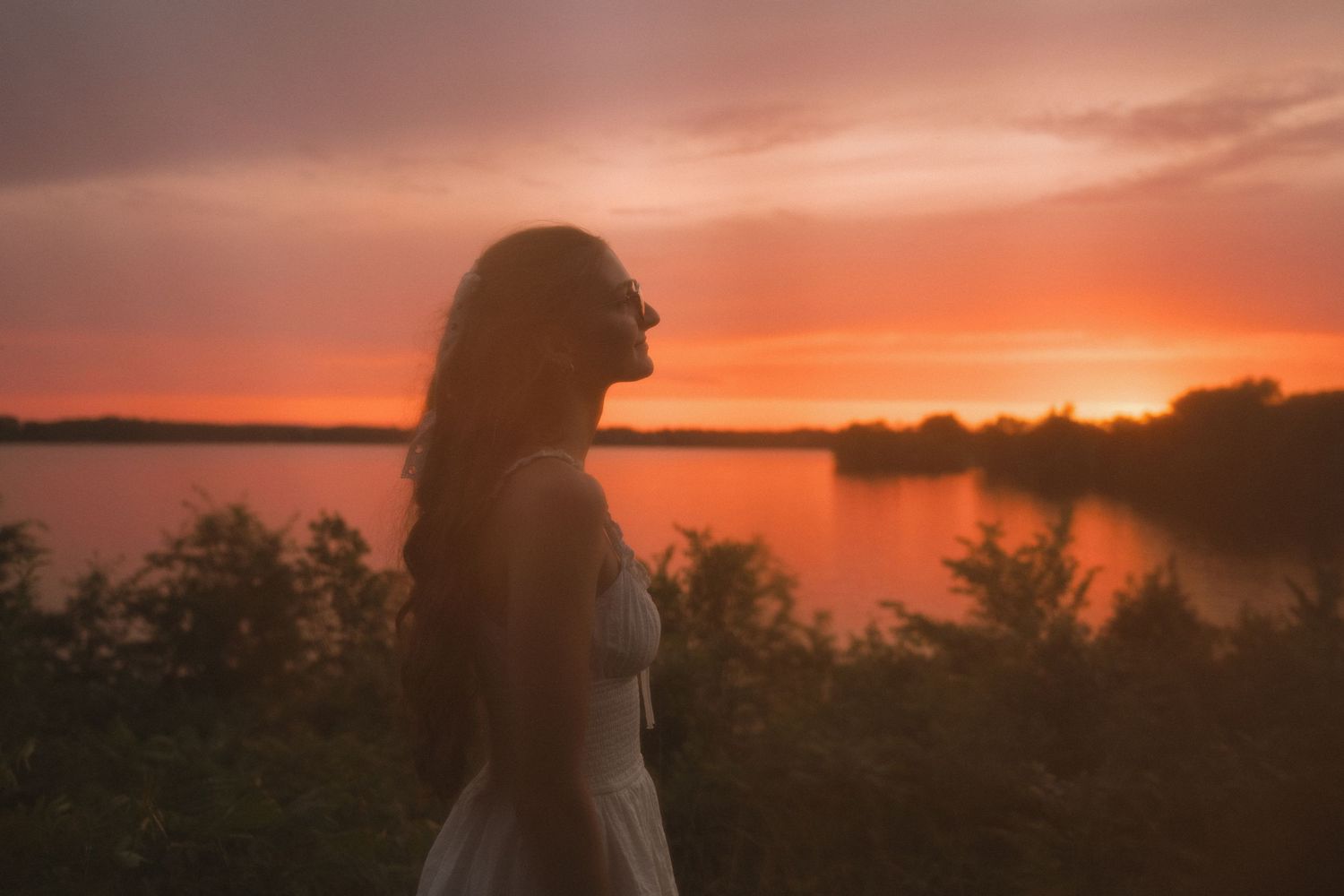 Young woman in white dress gazes at a vibrant orange and pink sunset over a calm lake surrounded by trees.