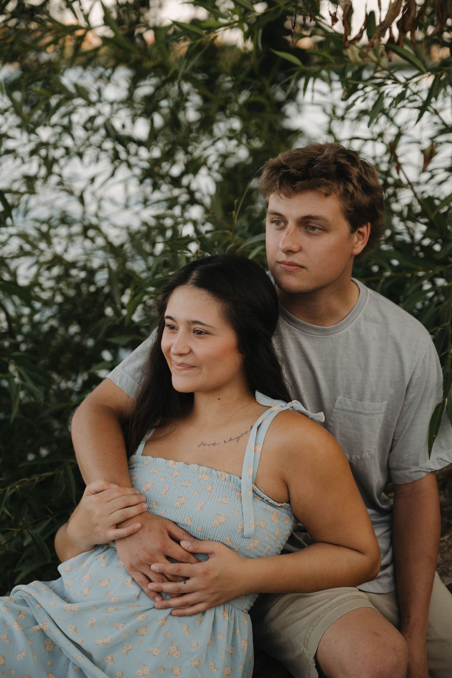 Young couple sitting together outdoors, man embracing woman in floral blue dress near green foliage.