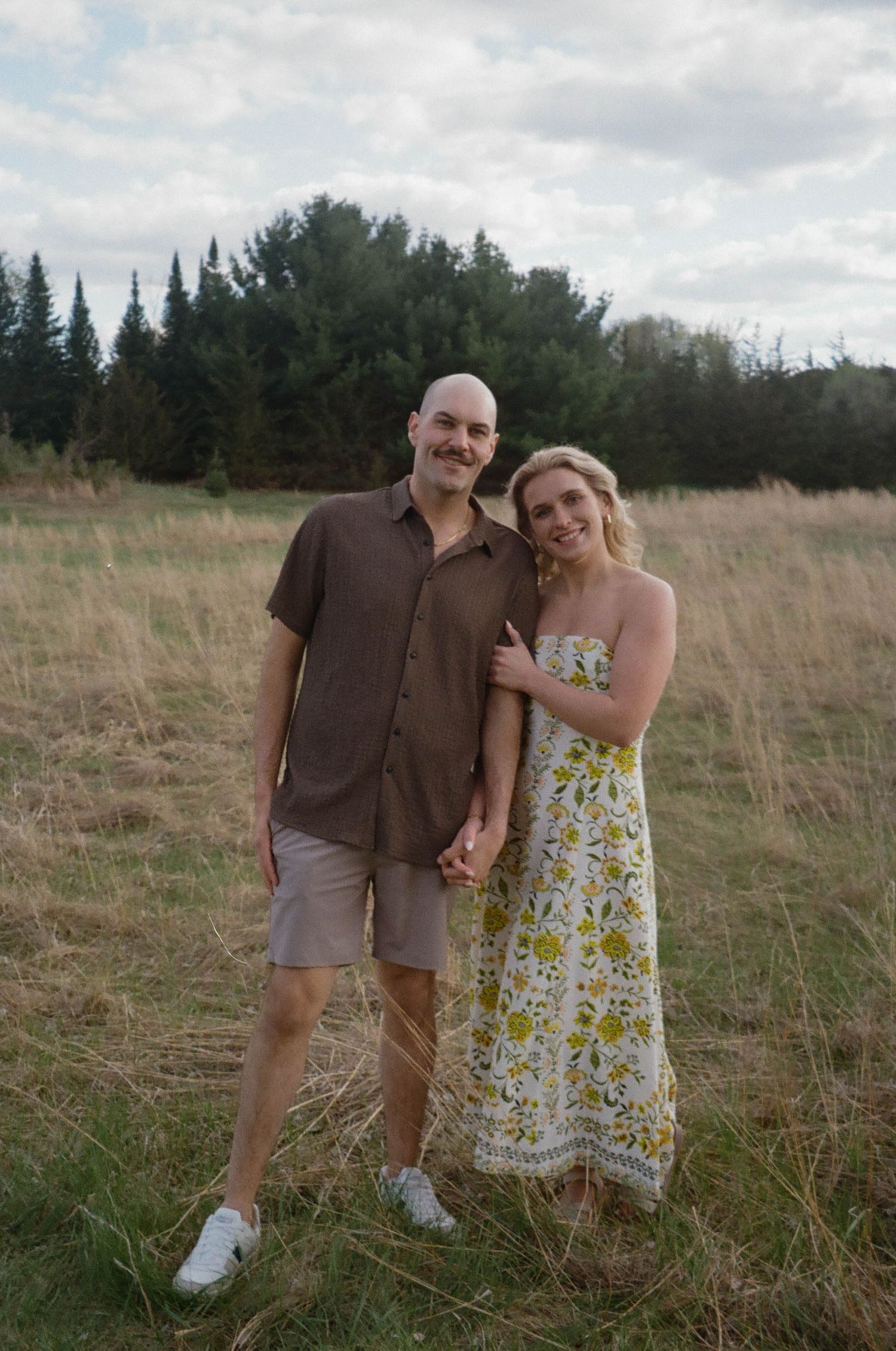 A smiling couple poses in a golden meadow with evergreen trees, man in brown shirt and shorts, woman in floral sundress.